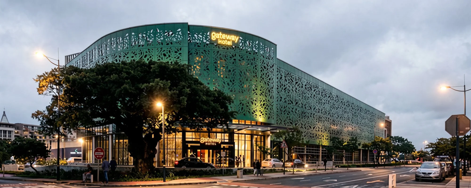 Modern hotel building with a decorative green facade and illuminated signs reading 'gateway Hotel' at dusk. The street in front has parked cars and people walking, with streetlights casting light.
