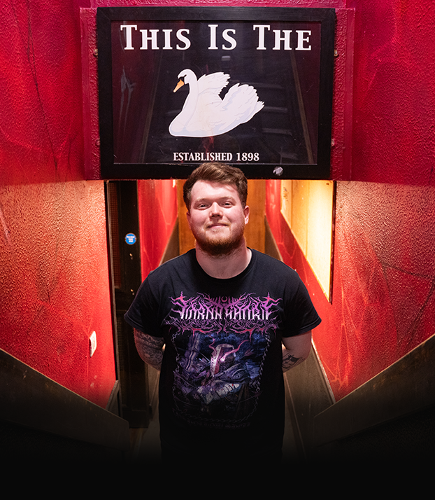 A young man with a beard and tattoos standing at the bottom of a stairway inside a building with red walls. Above him, a sign reads 'This Is The' with an illustration of a white swan and the text 'Established 1898.'