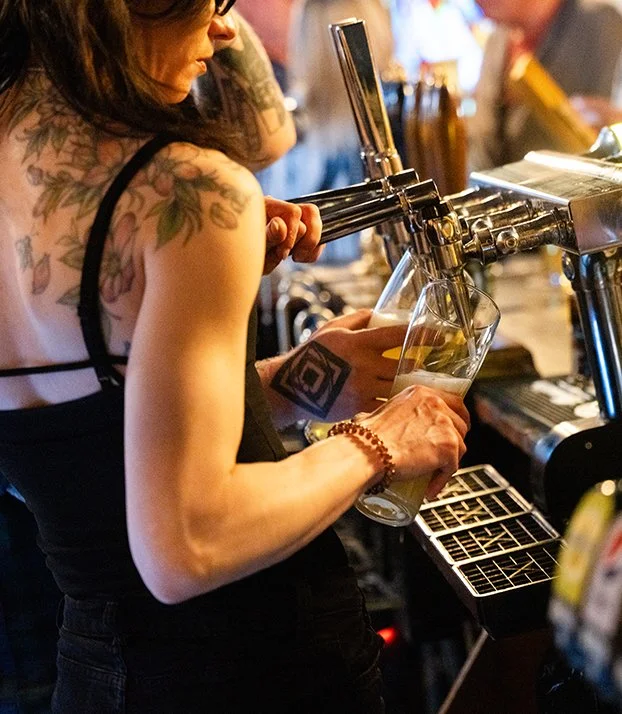 A woman with tattoos and a beaded bracelet pours draft beer from a tap into a glass at a bar.
