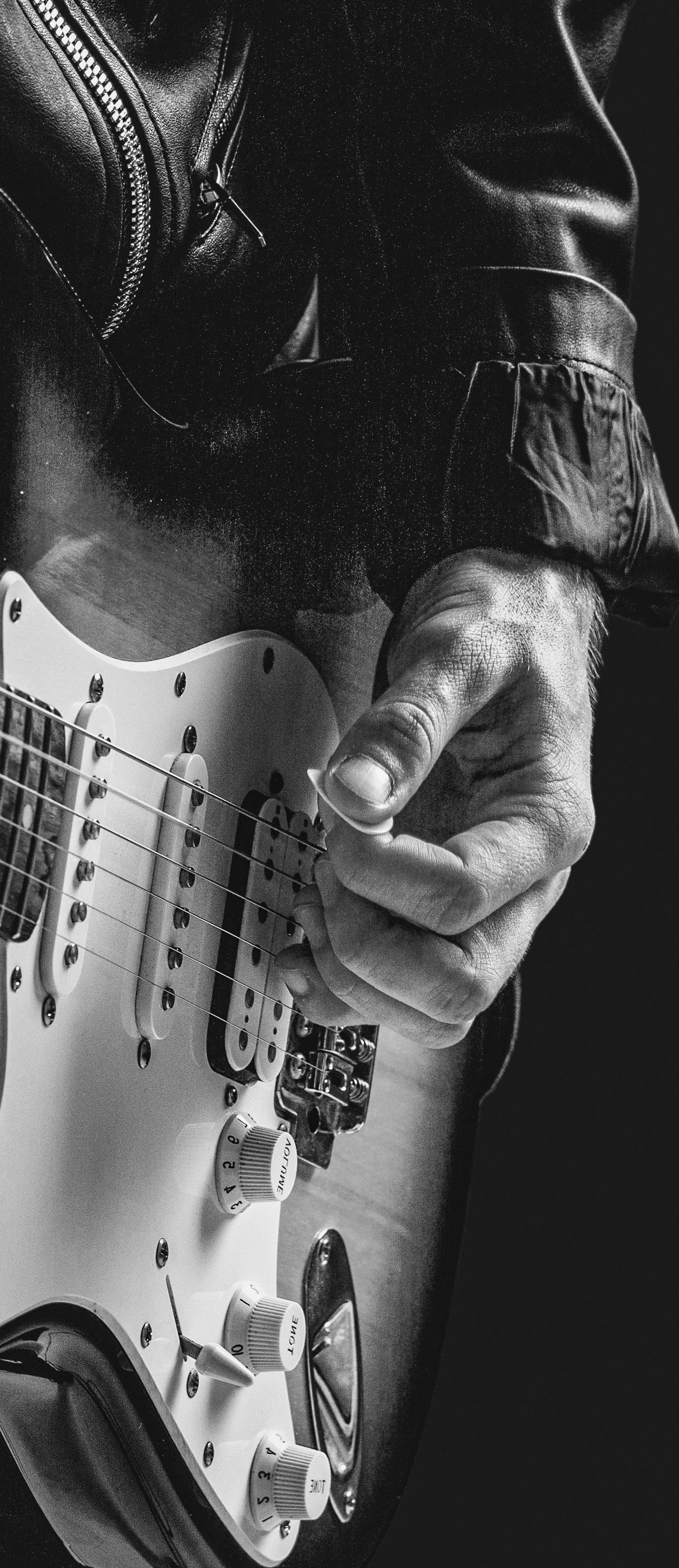 Close-up of a person's hand adjusting the tone control knob on an electric guitar, with part of their leather jacket visible.