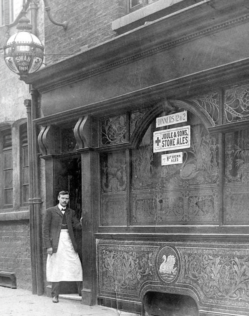 Man standing outside a historical pub with wood paneling and intricate decorative details, signs for dinners and stone ales, and a vintage lantern hanging from the building.