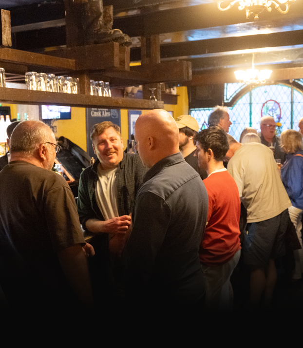 People socializing and chatting inside a dimly lit pub or bar, with wooden beams and stained-glass windows in the background.