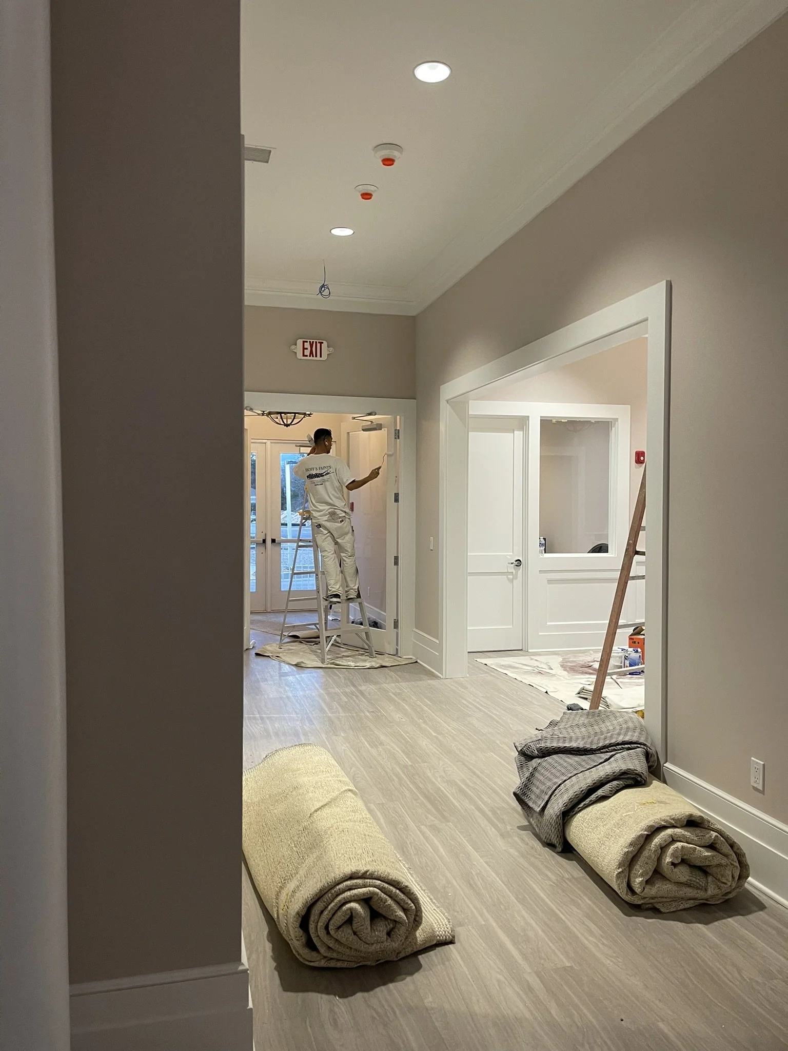 Interior view of a room under renovation with rolls of carpet on the floor, a worker on a ladder painting the wall, and construction tools around.
