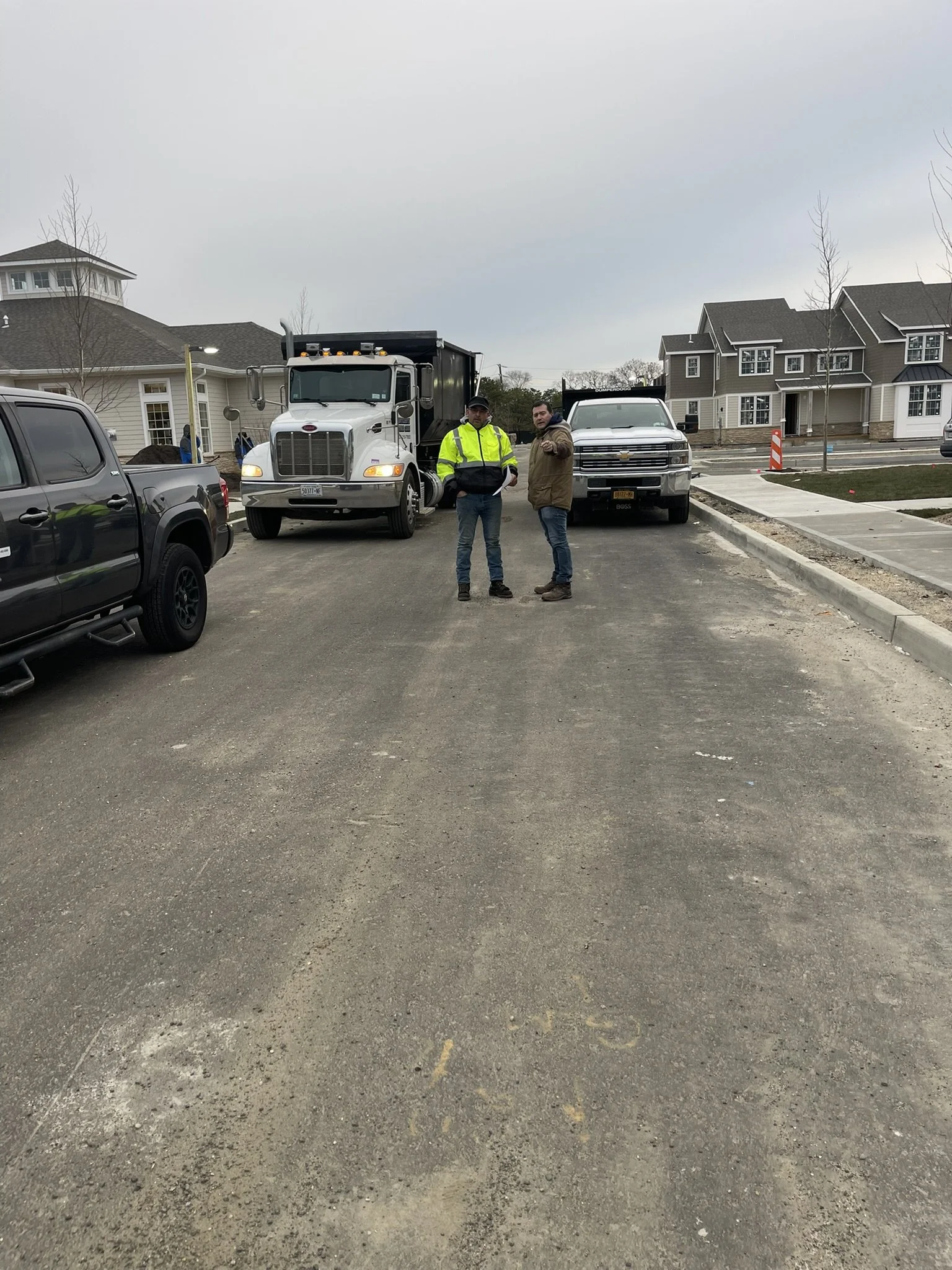 Construction site with two men standing on a gravel road, construction vehicles, and new houses in the background