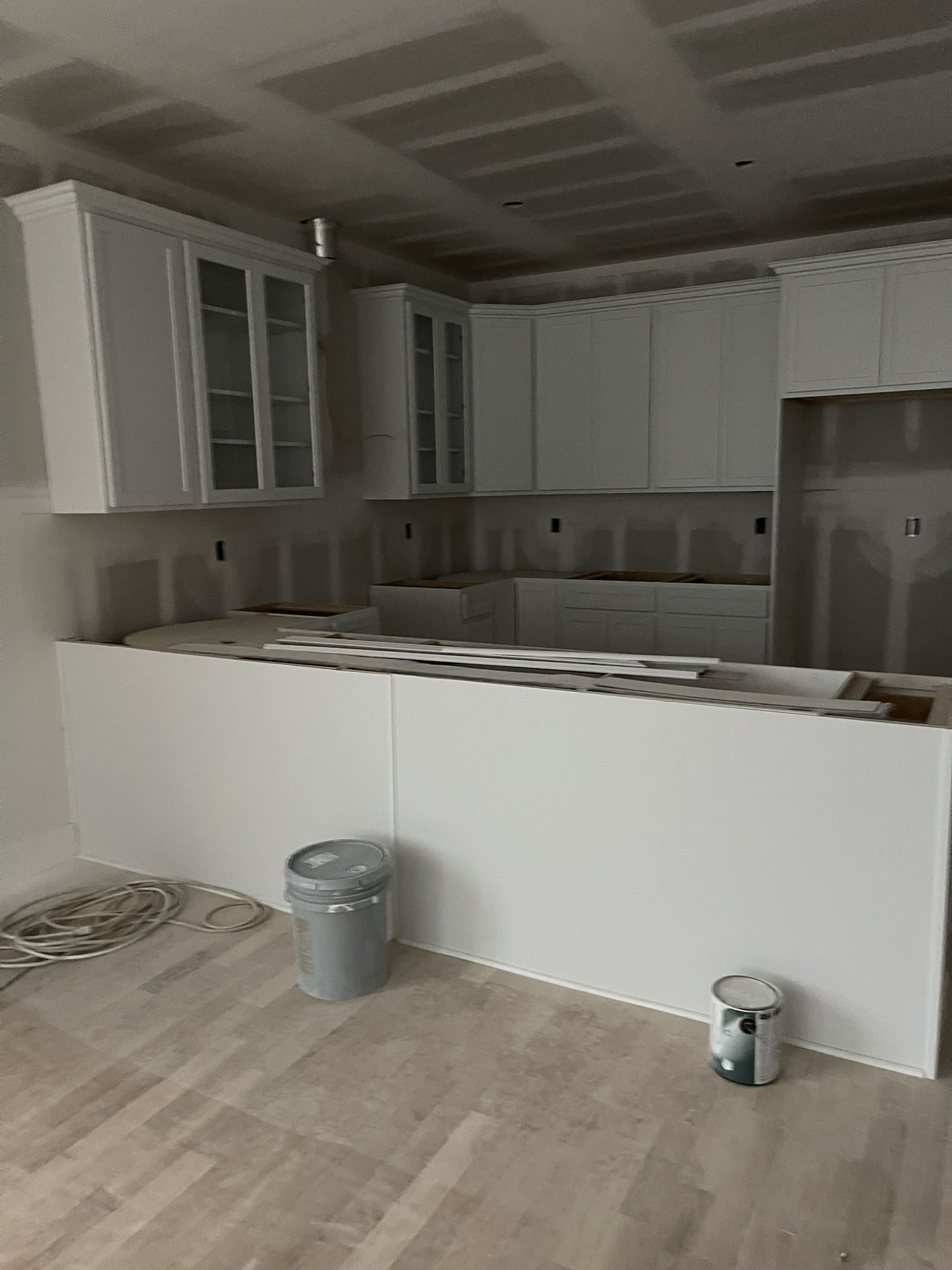 A kitchen under construction with white cabinets, a countertop, and a tiled floor. Construction materials and tools, including a bucket and some pipes, are scattered on the floor.