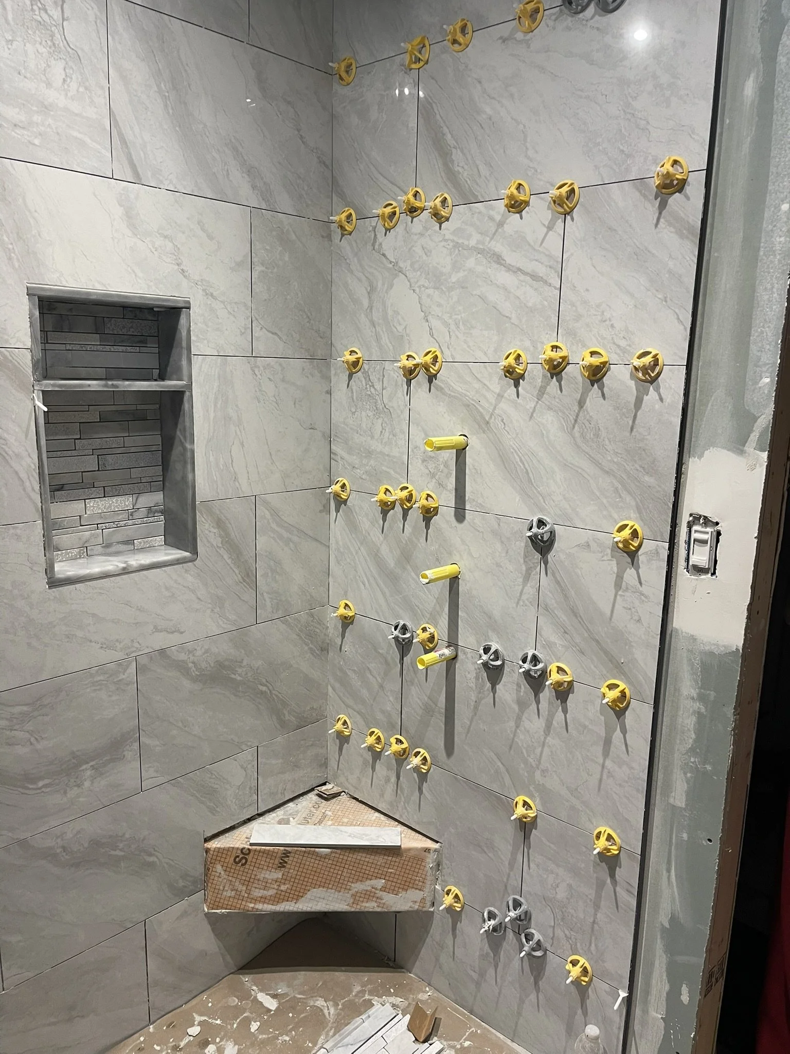 A bathroom shower wall being tiled with gray marble tiles. Yellow and gray tile spacers are attached between the tiles, and a small niche with gray bricks is built into the wall. Construction materials and debris are on the floor.
