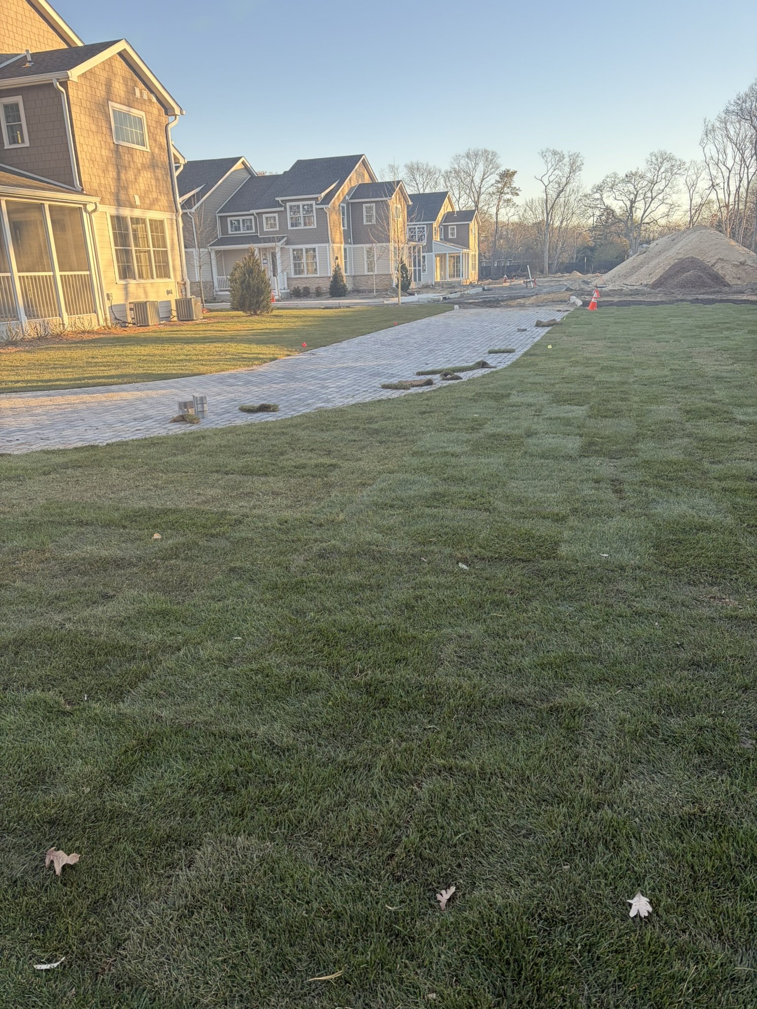 A suburban neighborhood with newly paved brick walkway, houses, and construction piles in the background, illuminated by warm late afternoon sunlight.