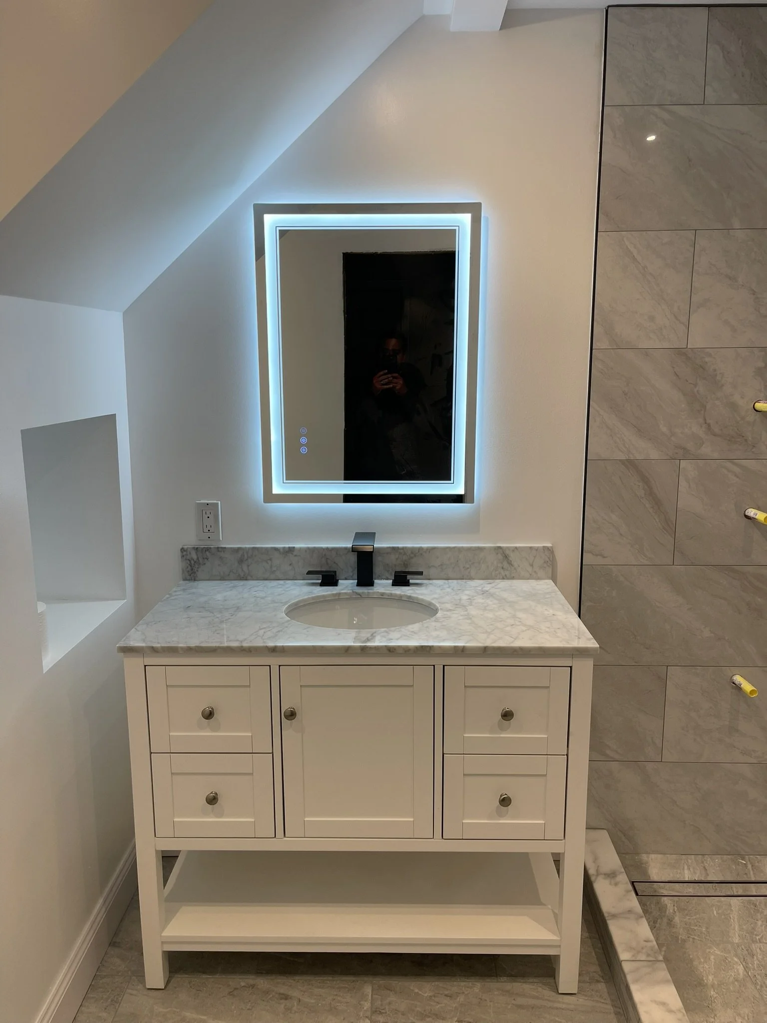 Bathroom vanity with white cabinet, marble countertop, and oval sink. Above it is a large rectangular mirror with LED lighting. To the right are marble tiles on the wall. The bathroom appears modern and well-lit.