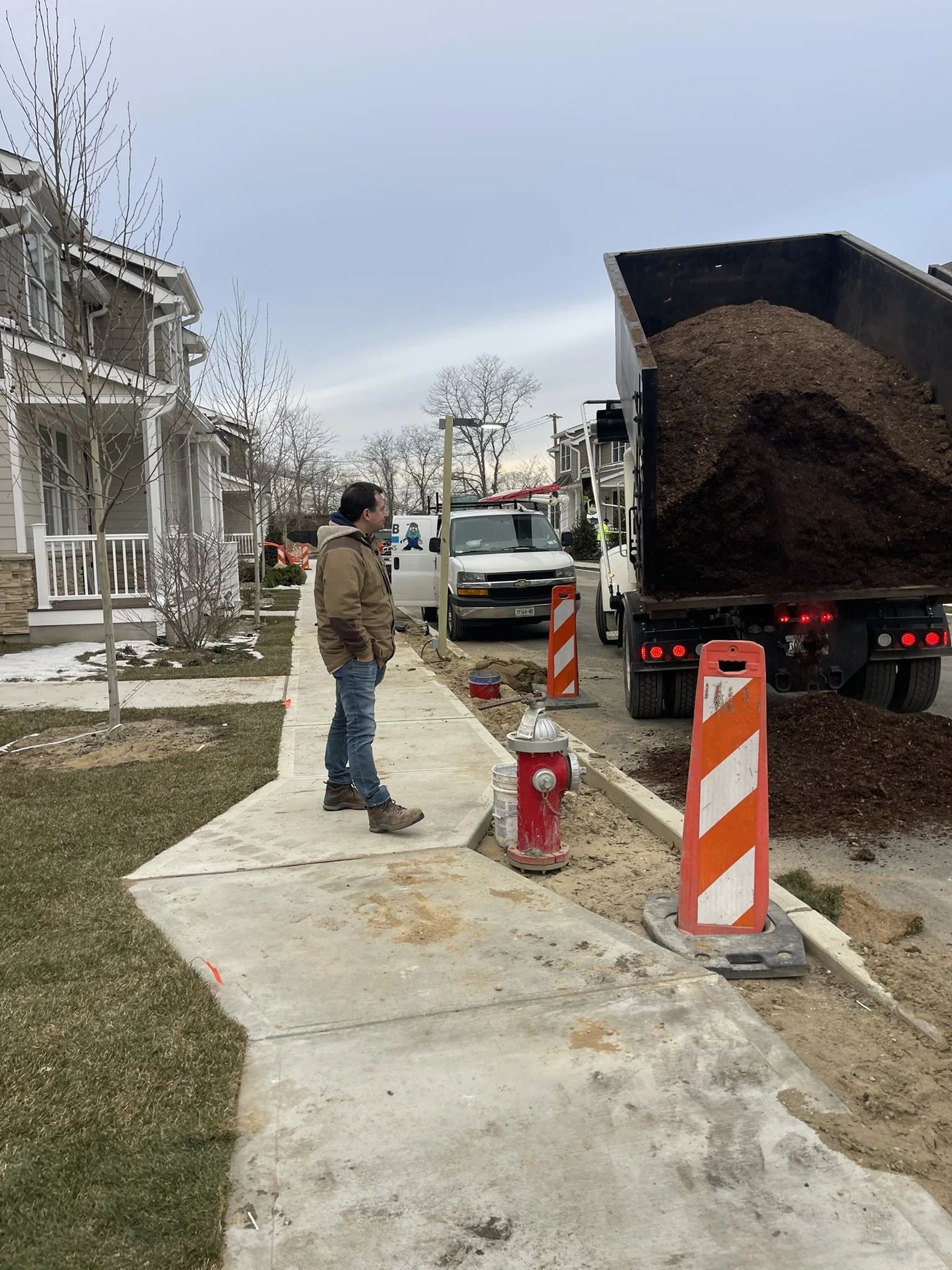 A man standing on a sidewalk while workers unload dirt from a large truck. The sidewalk is under construction, with traffic cones and barriers around it, and residential houses line the street.