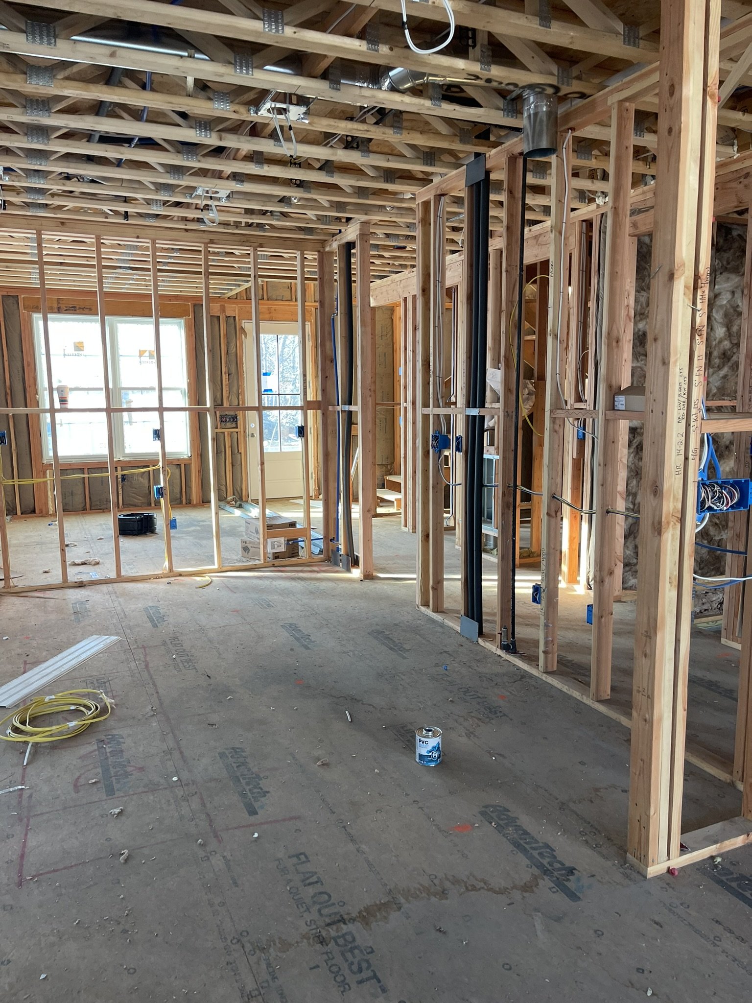 Interior of a house under construction showing wooden framing, ceiling trusses, and exposed wires. There's a window and an open doorway in the background, with construction materials and tools on the floor.
