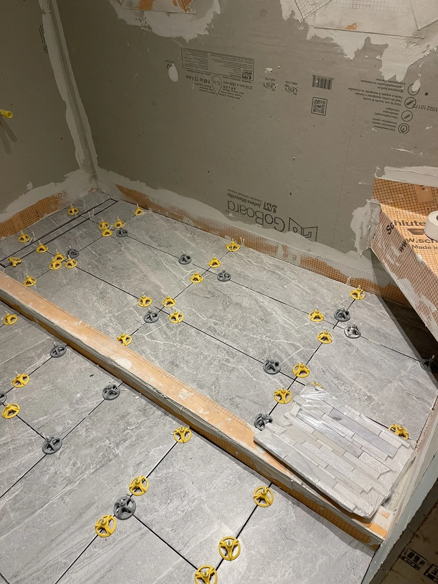 Building floor with tile installation in progress, showing yellow and gray tile spacers arranged in a grid pattern on the floor, with a wall in the background covered in drywall and construction materials.