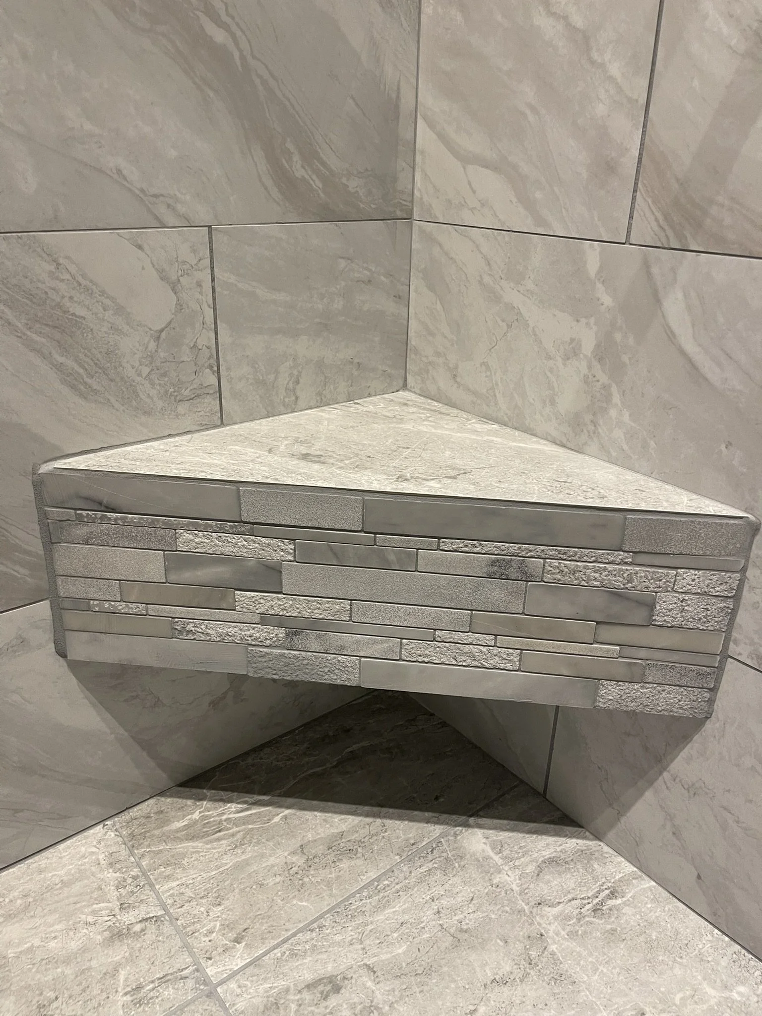 Corner of a tiled bathroom wall with large marble tiles above and small mosaic tiles below, featuring shades of gray and beige.