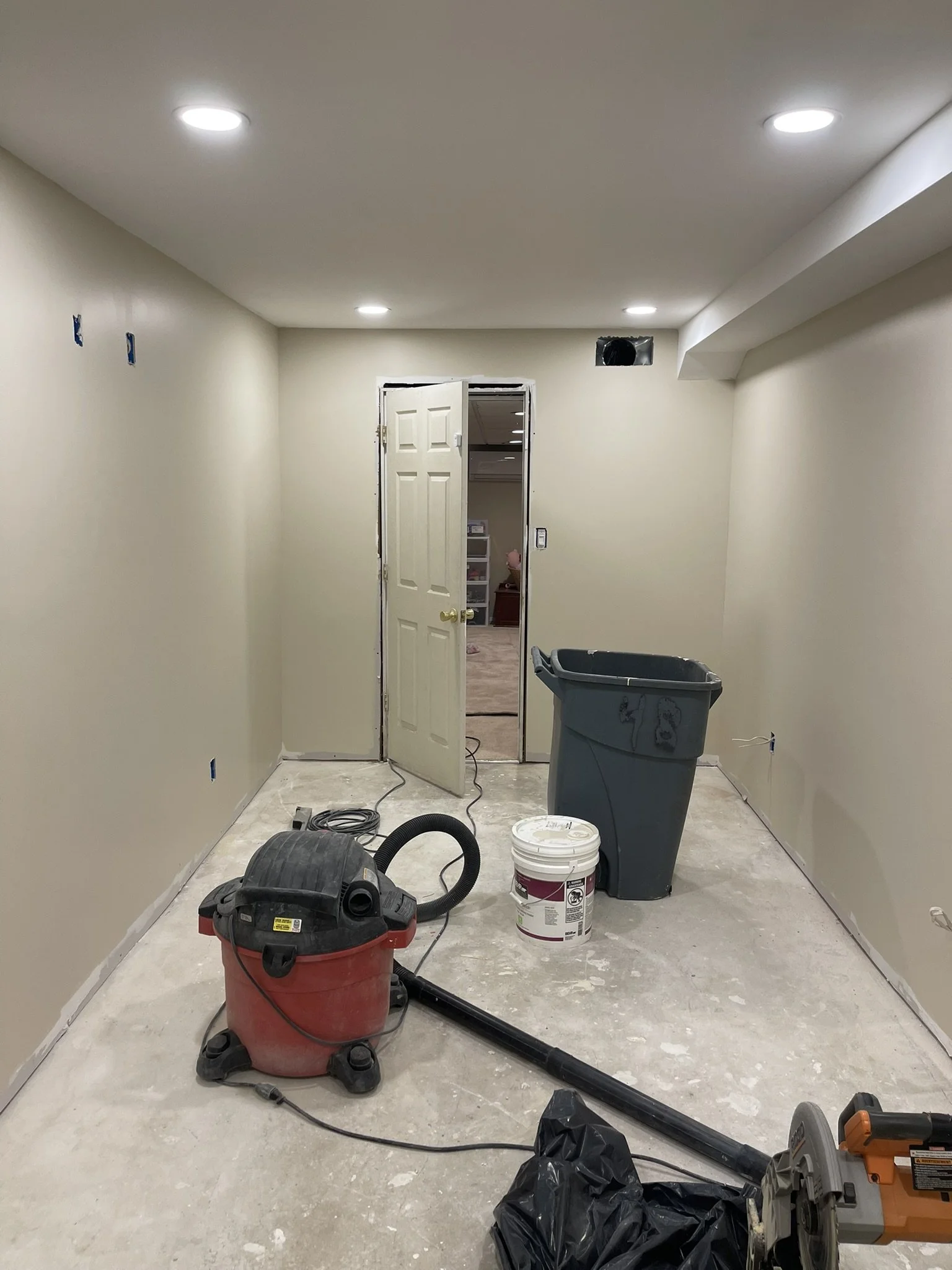 An unfinished room with beige walls and a ceiling with recessed lighting. There is construction equipment and tools on the floor, including a wet/dry vacuum, a trash bin, a bucket, a toolbox, and other power tools, indicating ongoing renovation work.