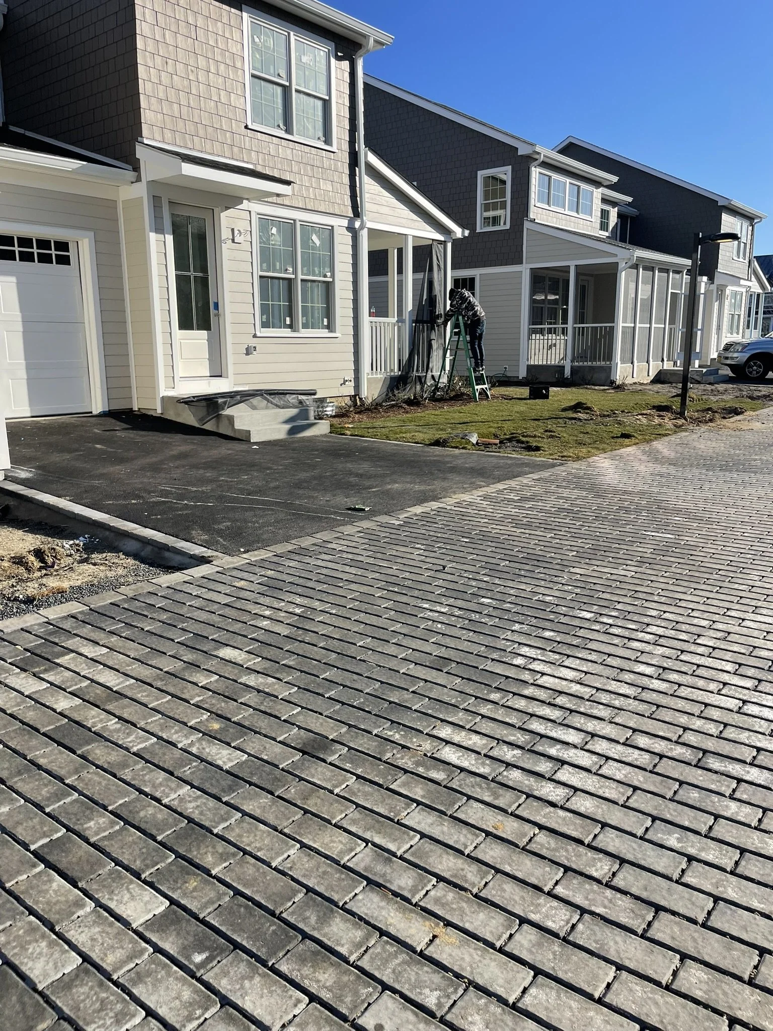 Newly paved black asphalt driveway in front of a row of modern townhouses, with a paved sidewalk and a lamppost under a clear blue sky.