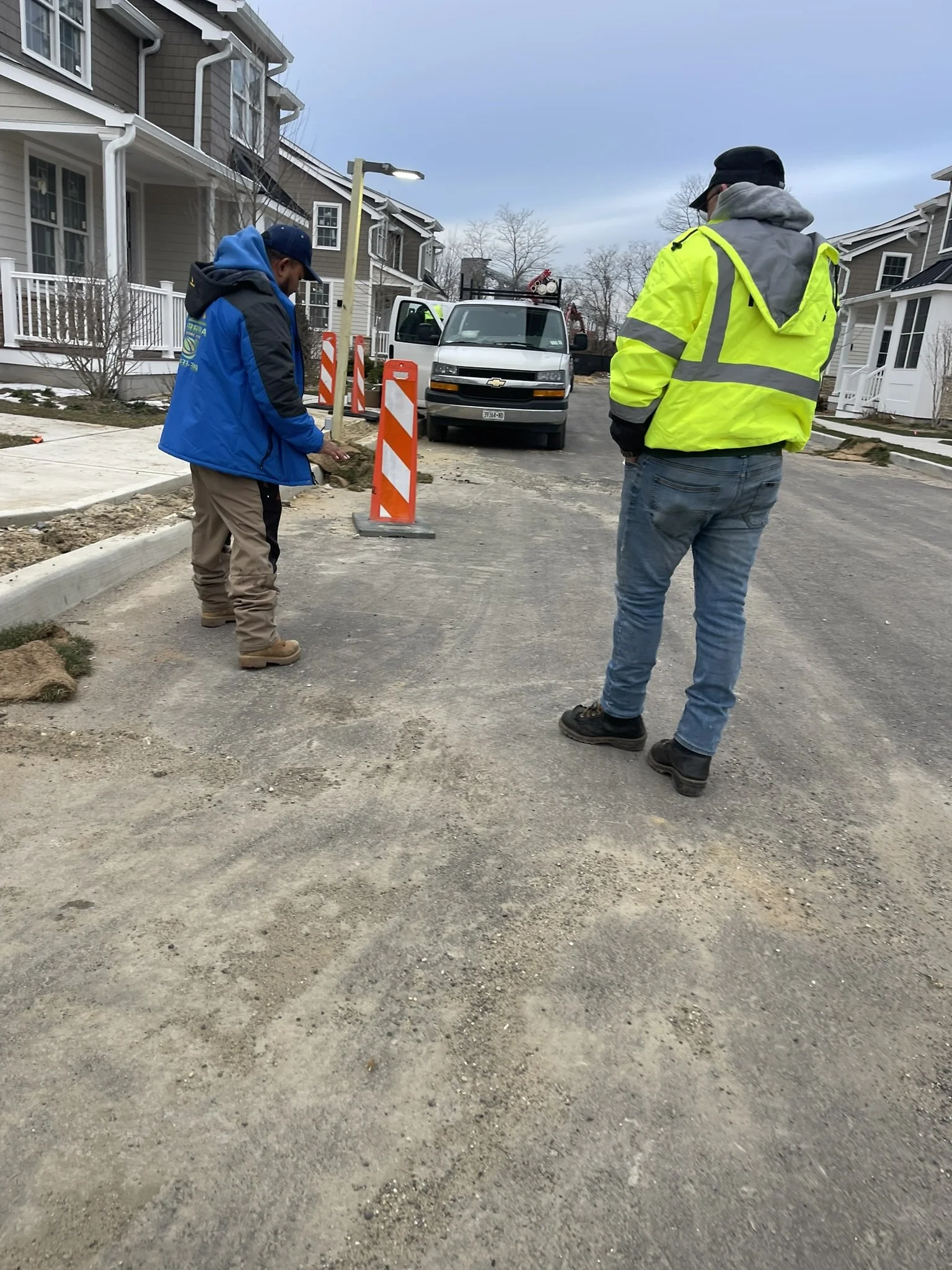 Two workers wearing safety gear inspecting a construction site on a residential street, with orange traffic barriers and a parked Chevrolet van.