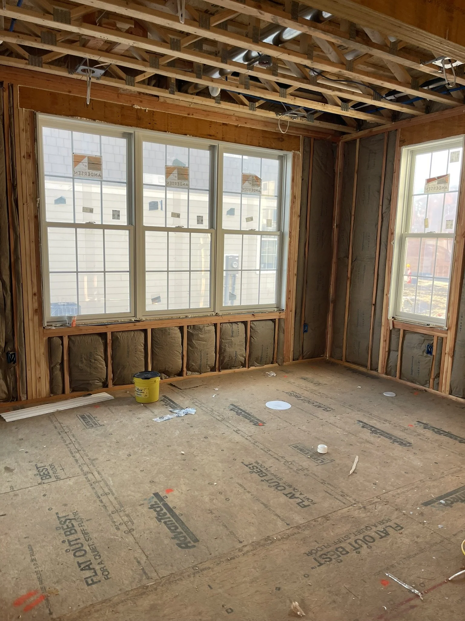 Room under construction with exposed wooden framing, insulation in the walls, and new windows installed. The floor is covered with construction material and tools.