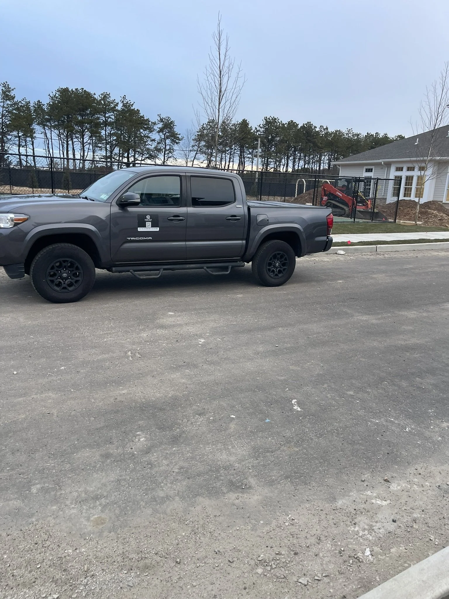 A gray Toyota Tacoma pickup truck parked on a dirt lot next to a sidewalk, with a house under construction and a fenced backyard in the background.