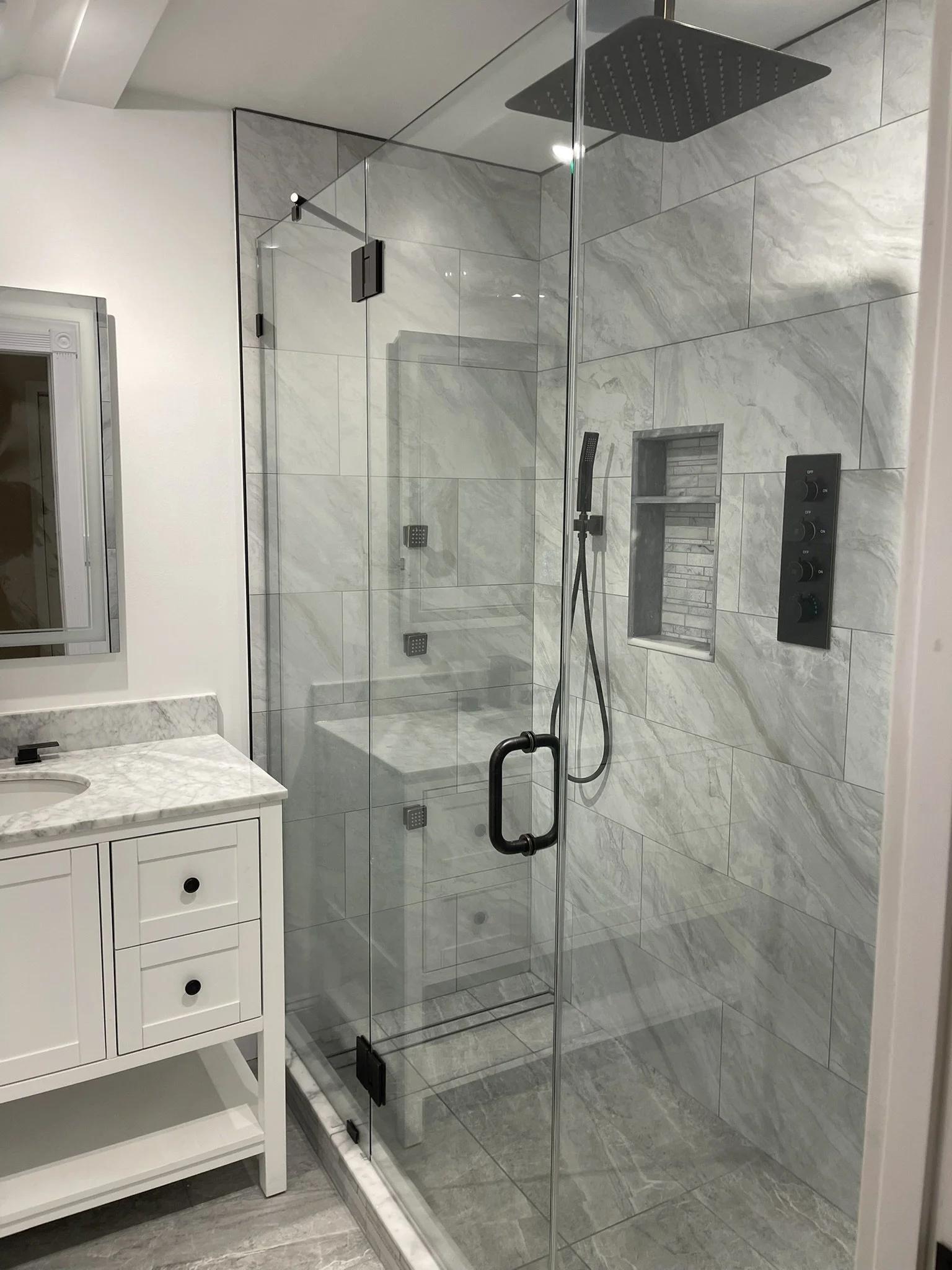 Modern bathroom with a glass-enclosed walk-in shower featuring a rain showerhead and a handheld shower, tiled in gray marble. A built-in niche with shelves is visible in the shower wall. Part of a white vanity with a marble countertop and a small mir