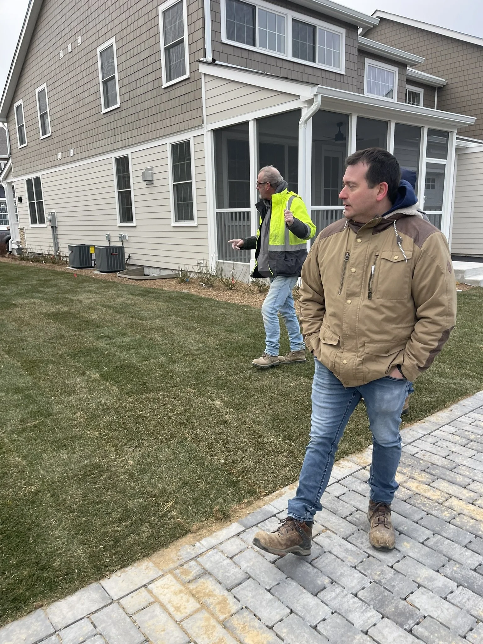 Two men walking outside near a house with a screened porch. One man wears a yellow safety jacket; the other wears a tan jacket and jeans. Paved walkway and grass in the yard.