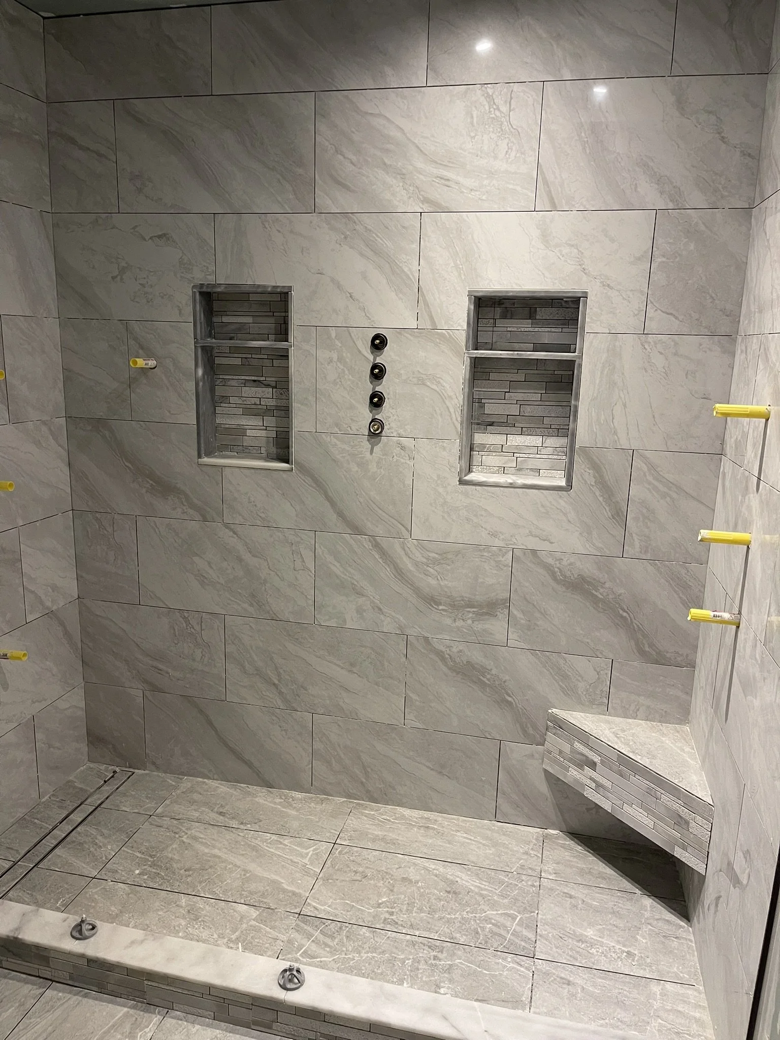 A partially constructed bathroom with beige marble tiled walls and floor, built-in shower niches with gray tile backsplashes, and yellow wall anchors on the walls. Showers drain on the floor, and there is a small marble bench in the corner.