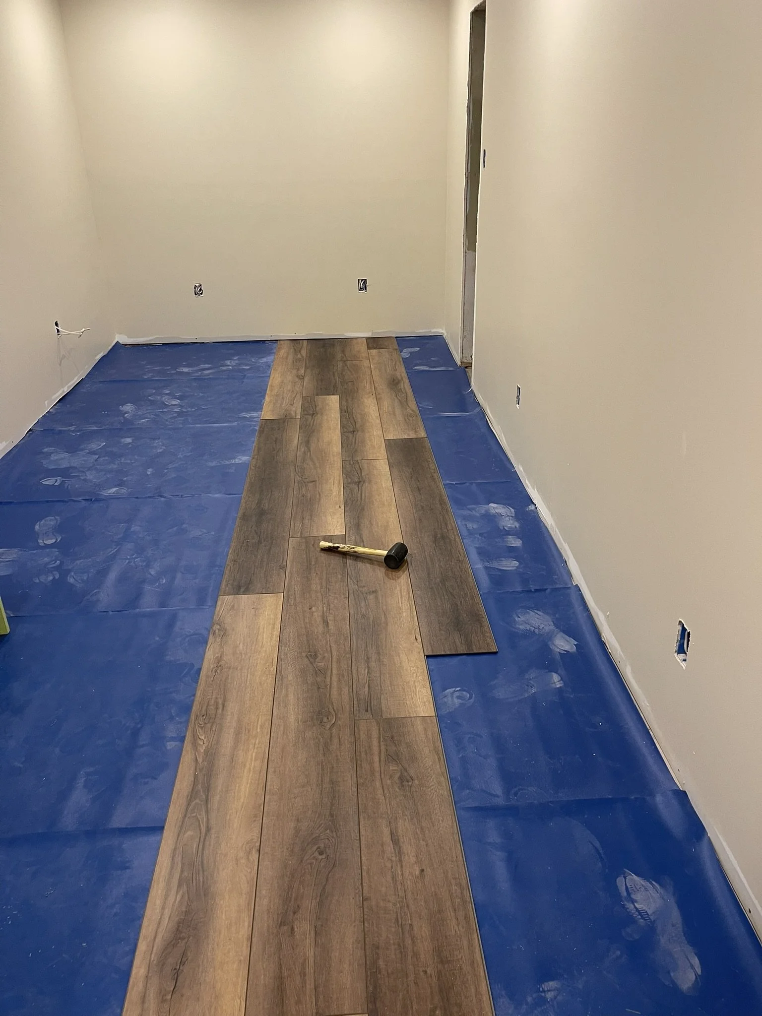 Room under renovation with beige walls, partially installed wood flooring, blue painter's plastic on the floor, and a mallet resting on the new flooring.