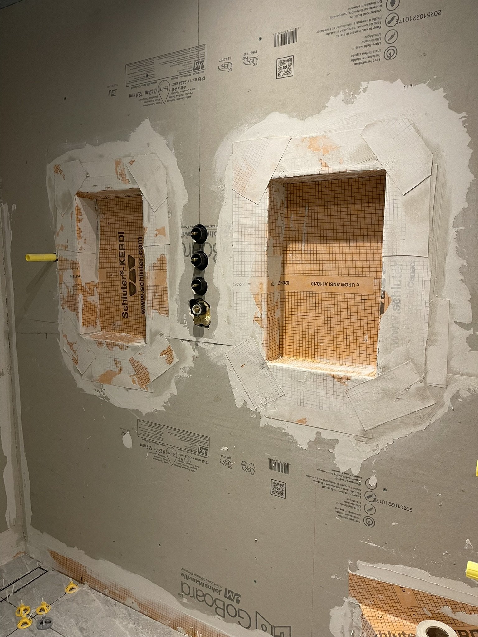 Bathroom or kitchen wall under construction with drywall and cutouts for fixtures, electrical wiring visible, and some yellow and gray plumbing fixtures on the floor.