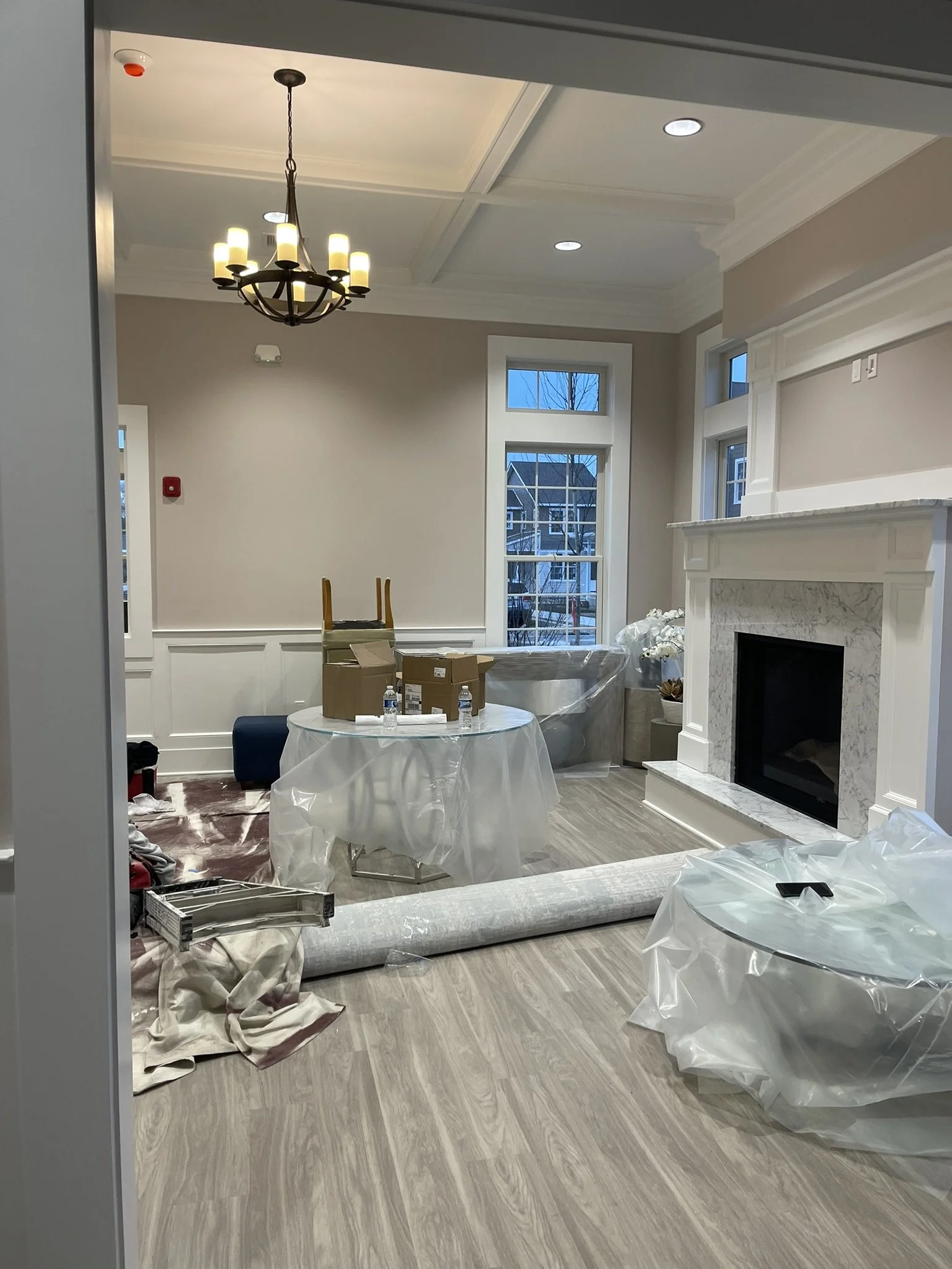 Interior of a living room under renovation, with plastic covers on furniture, cardboard boxes on a round table, and construction materials on the floor, a fireplace with a marble surround, large windows showing neighboring houses, and a chandelier ha