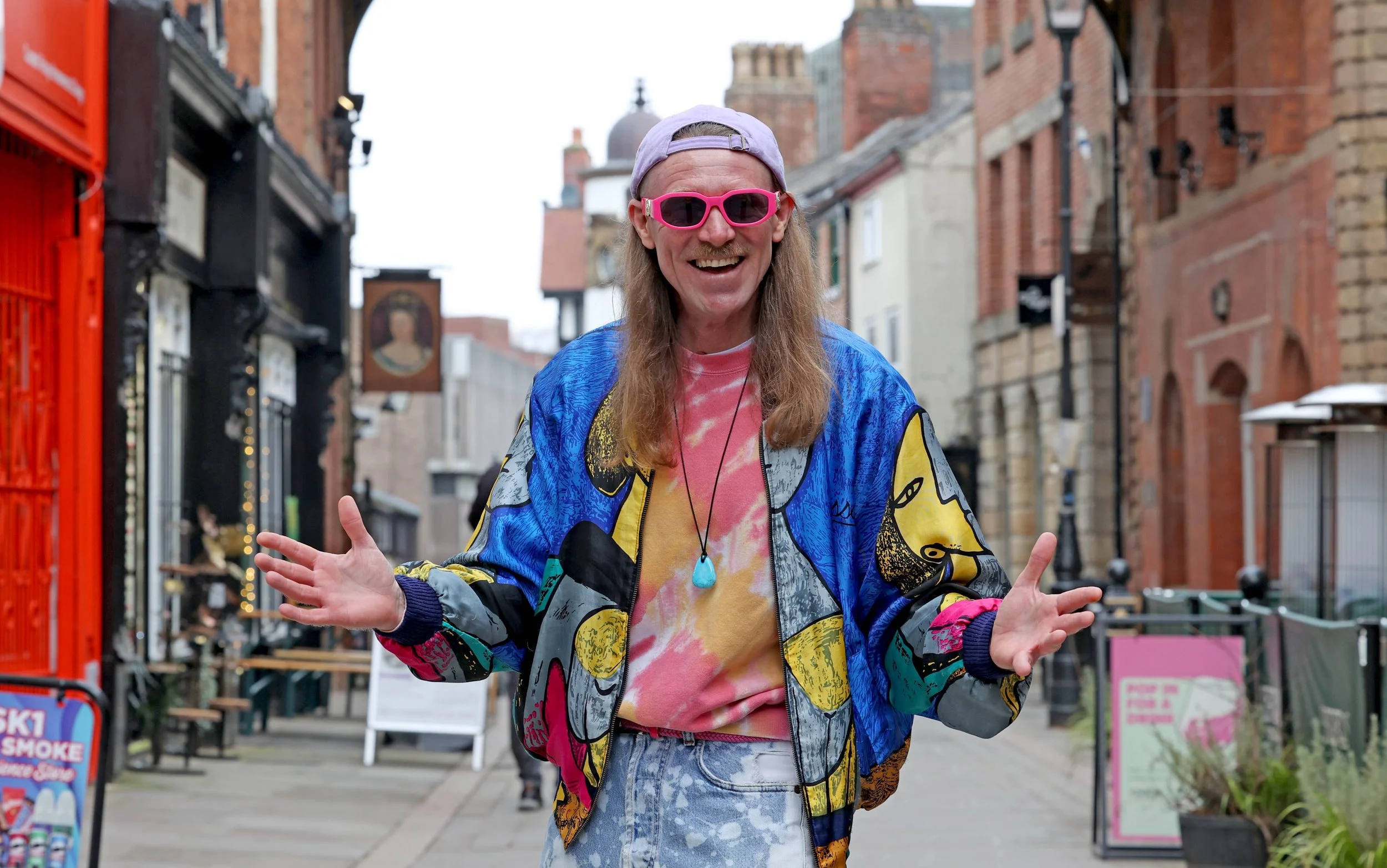 A man with long hair, wearing a colorful jacket, pink sunglasses, a tie-dye shirt, and a purple cap, smiling and gesturing with his hands on a city street.