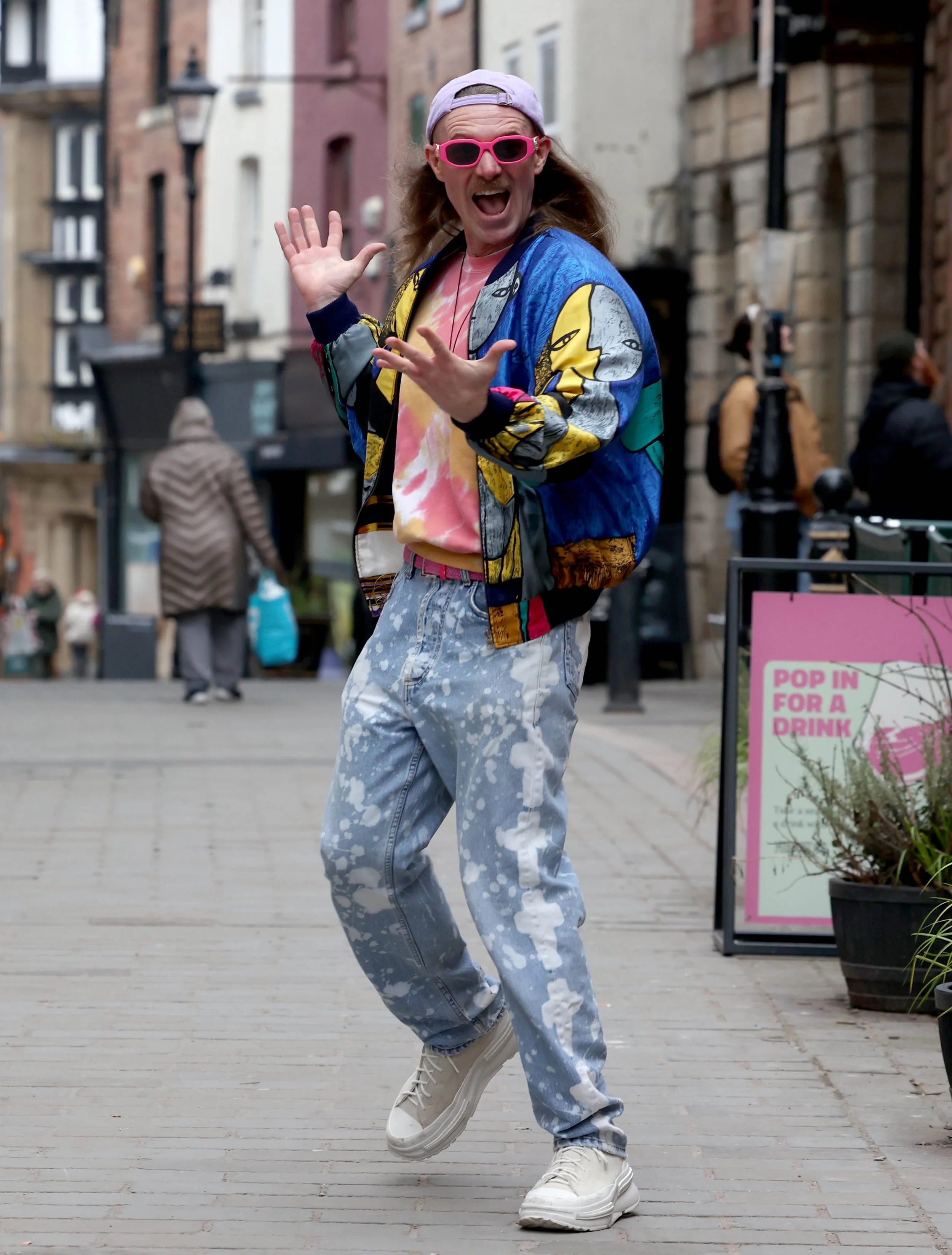 A person with long hair, pink sunglasses, and a purple cap, wearing a colorful jacket, tie-dye shirt, and paint-splattered jeans, dancing on a city street.