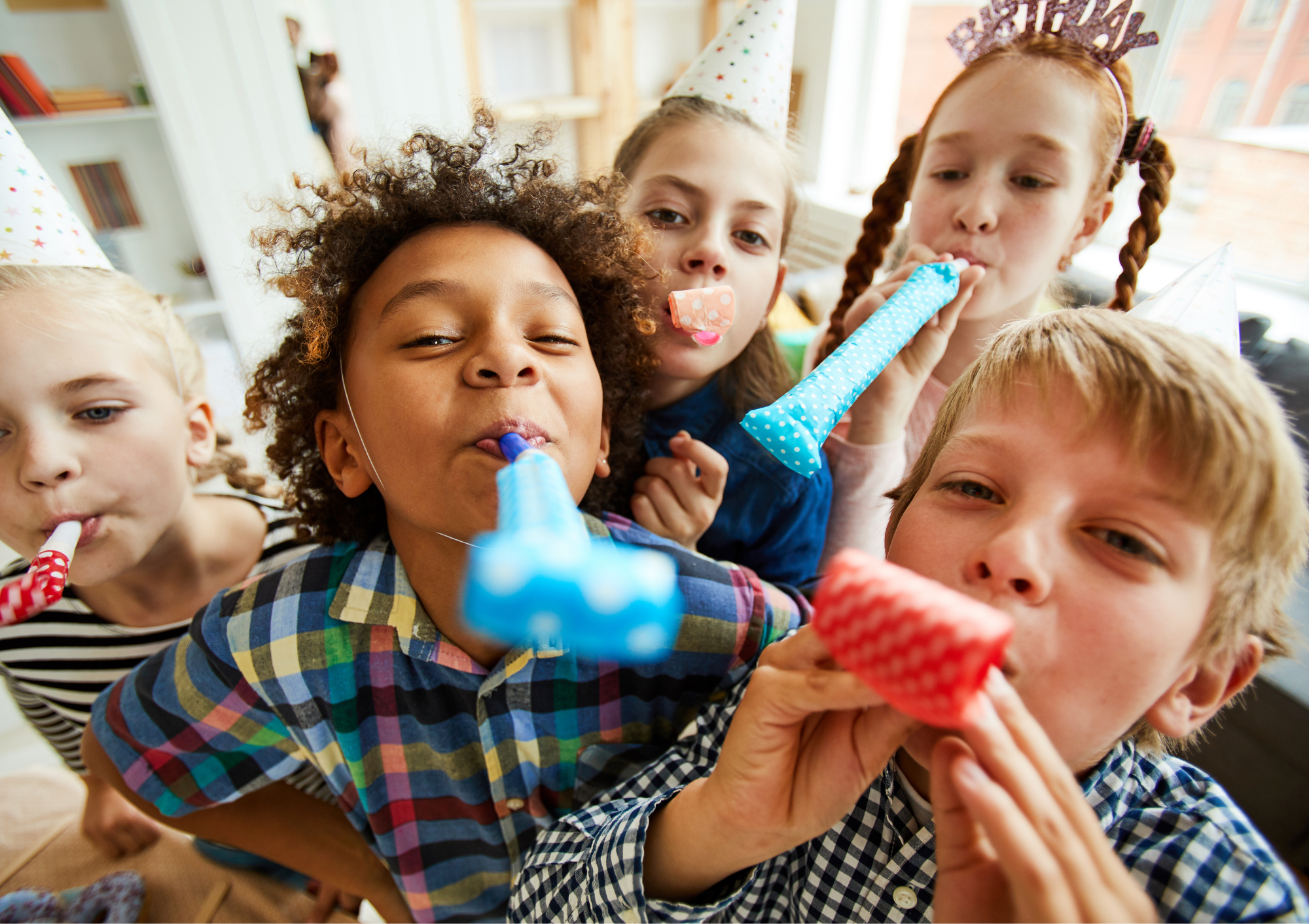 Group of children celebrating a party, wearing party hats, with some blowing party blowers and others holding items, in a bright room with windows.