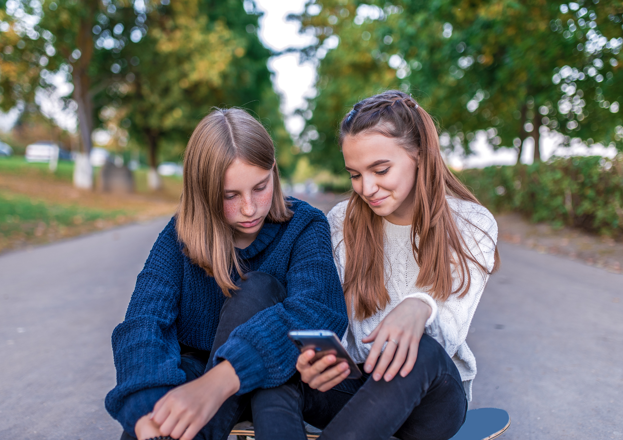 Two girls sitting outdoors on a park bench, looking at a smartphone together, with trees and cars in the background.