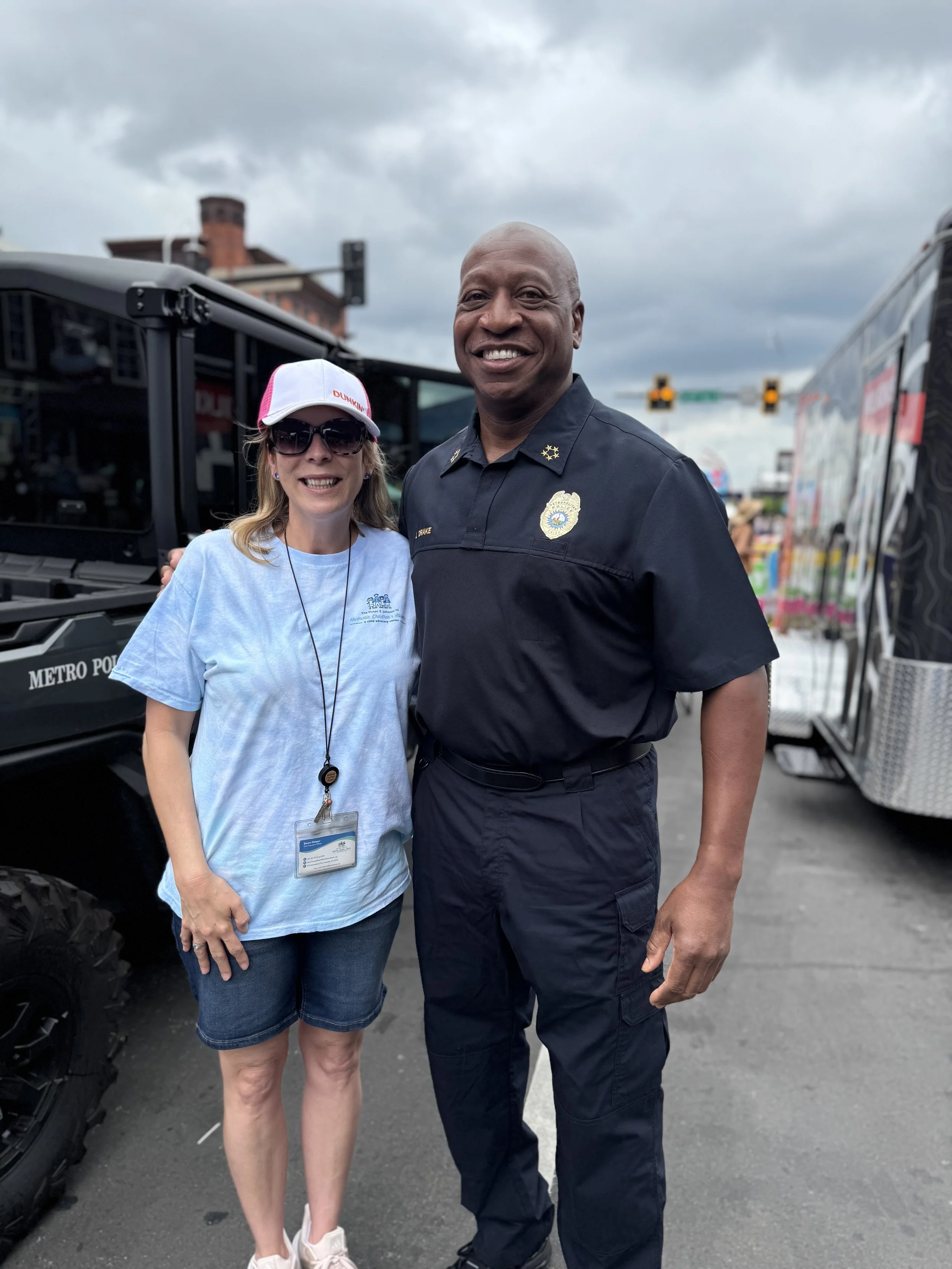 A woman wearing sunglasses, a white and pink Dunkin' Donuts cap, and a light blue t-shirt standing next to a police officer in uniform. They are outdoors on a cloudy day, with a police vehicle and a vending truck in the background.