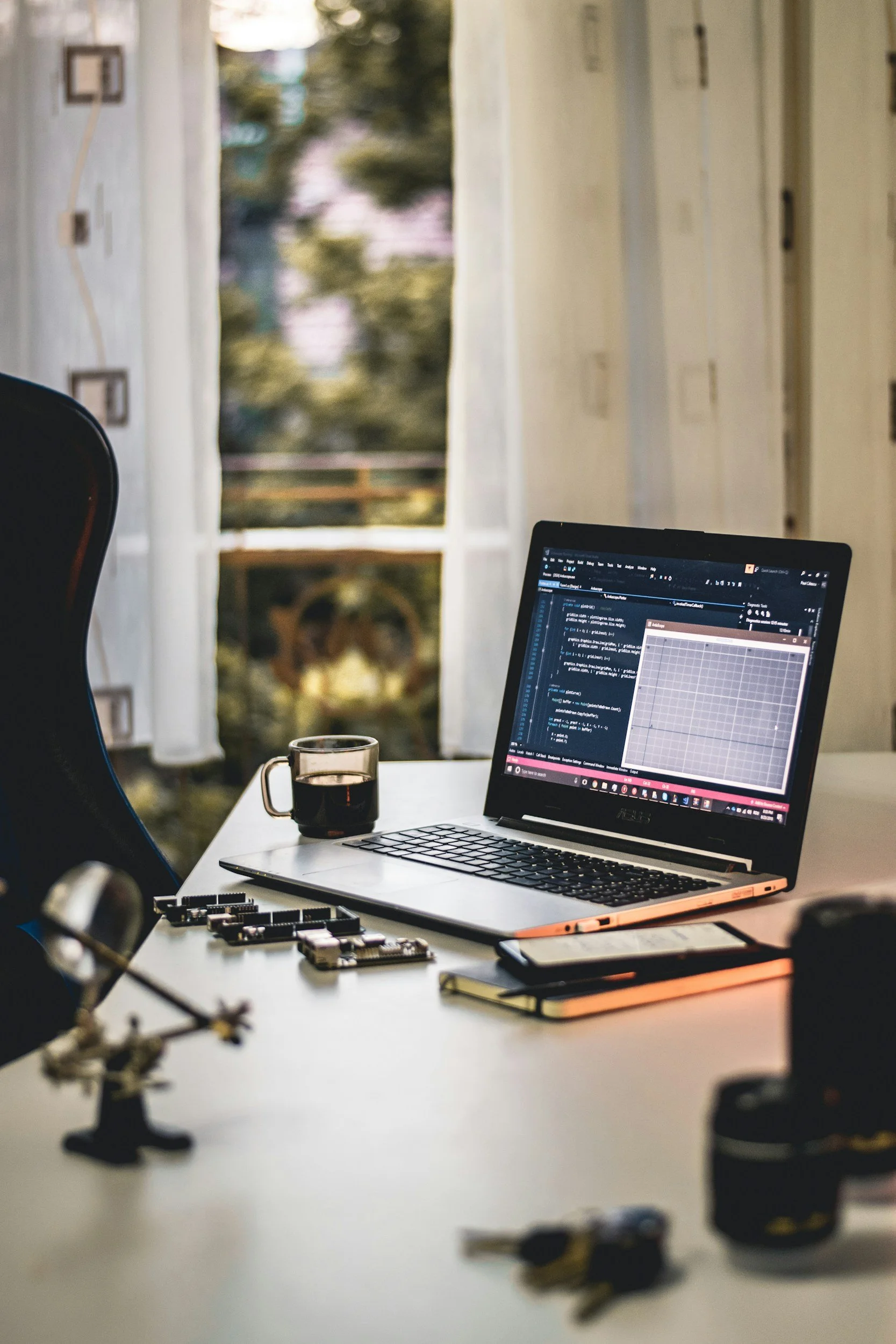 Work desk with a laptop showing a programming interface, a cup of coffee, a notebook, a smartphone, and various electronic components on a white table near an open window with curtains, sunlight streaming in.