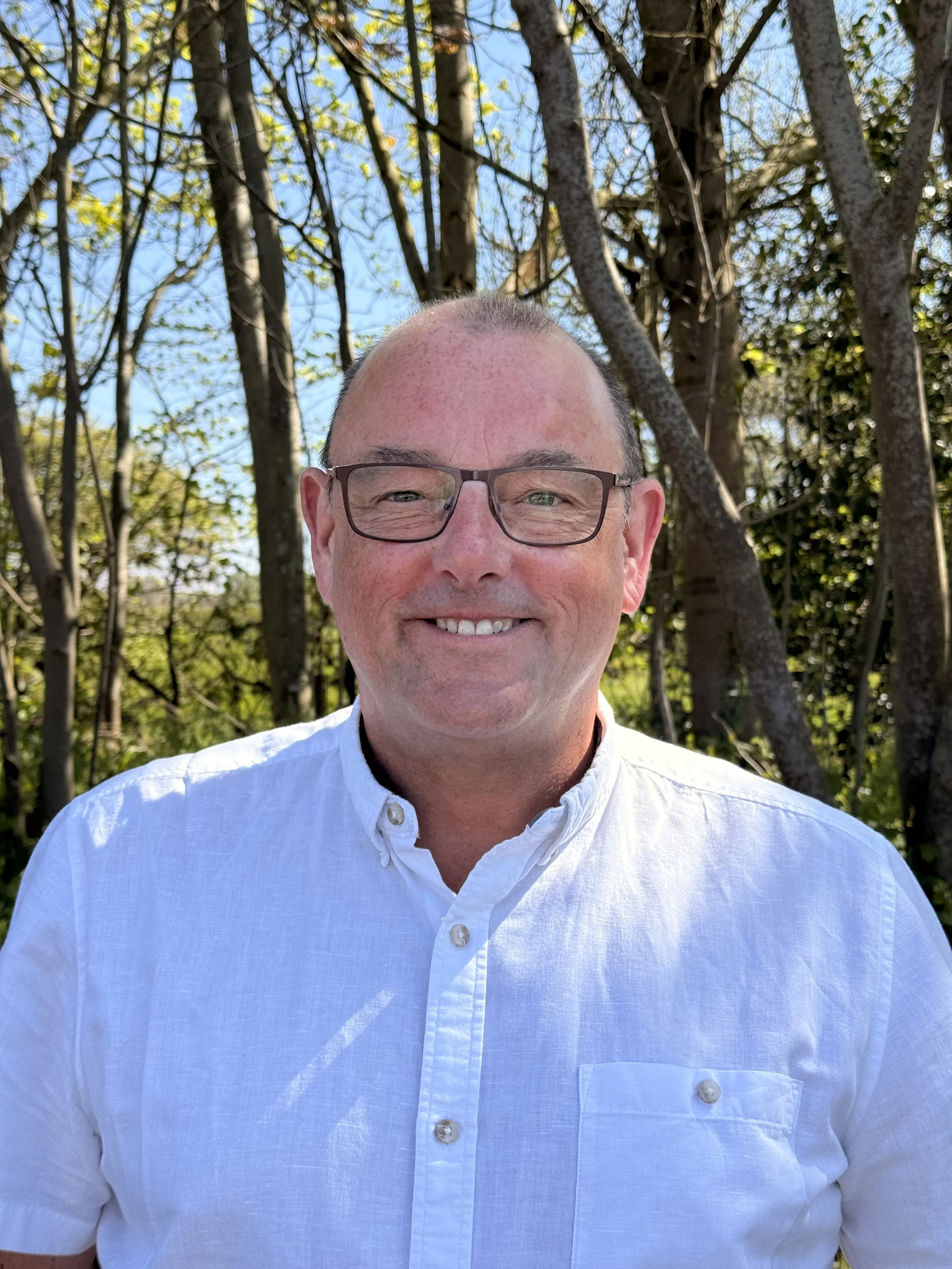A smiling man with glasses dressed in a white shirt standing outdoors in front of trees with green leaves and a blue sky.