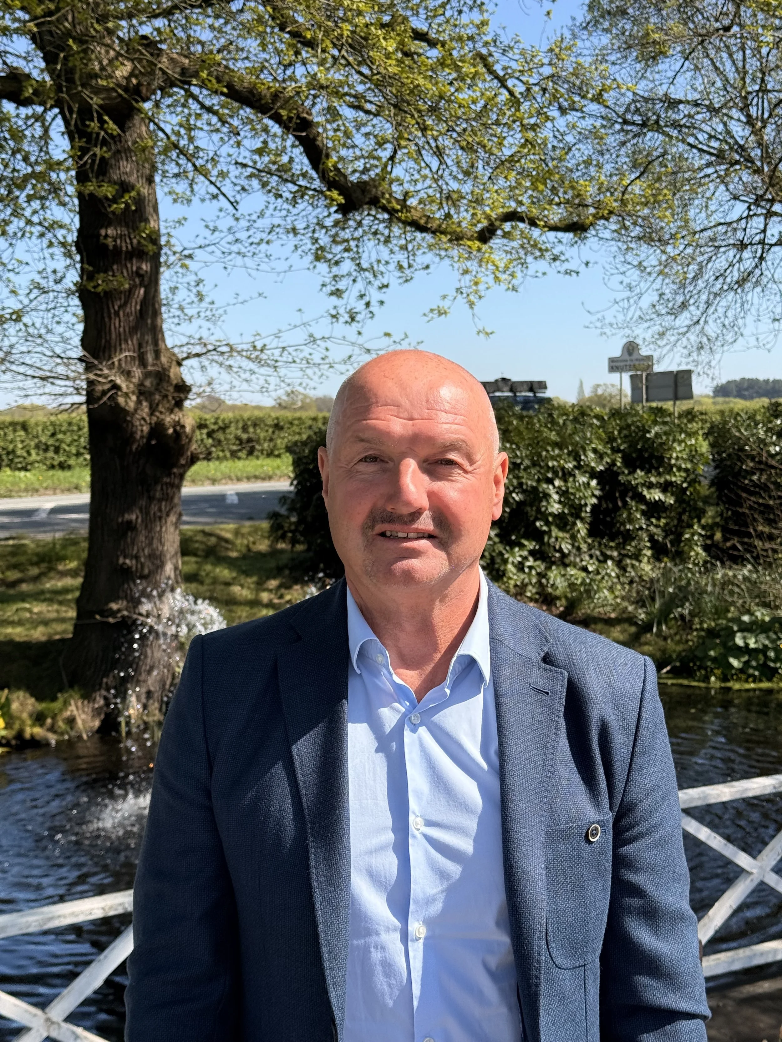 A man in a suit and white shirt taking a selfie outdoors near water with trees and greenery in the background.