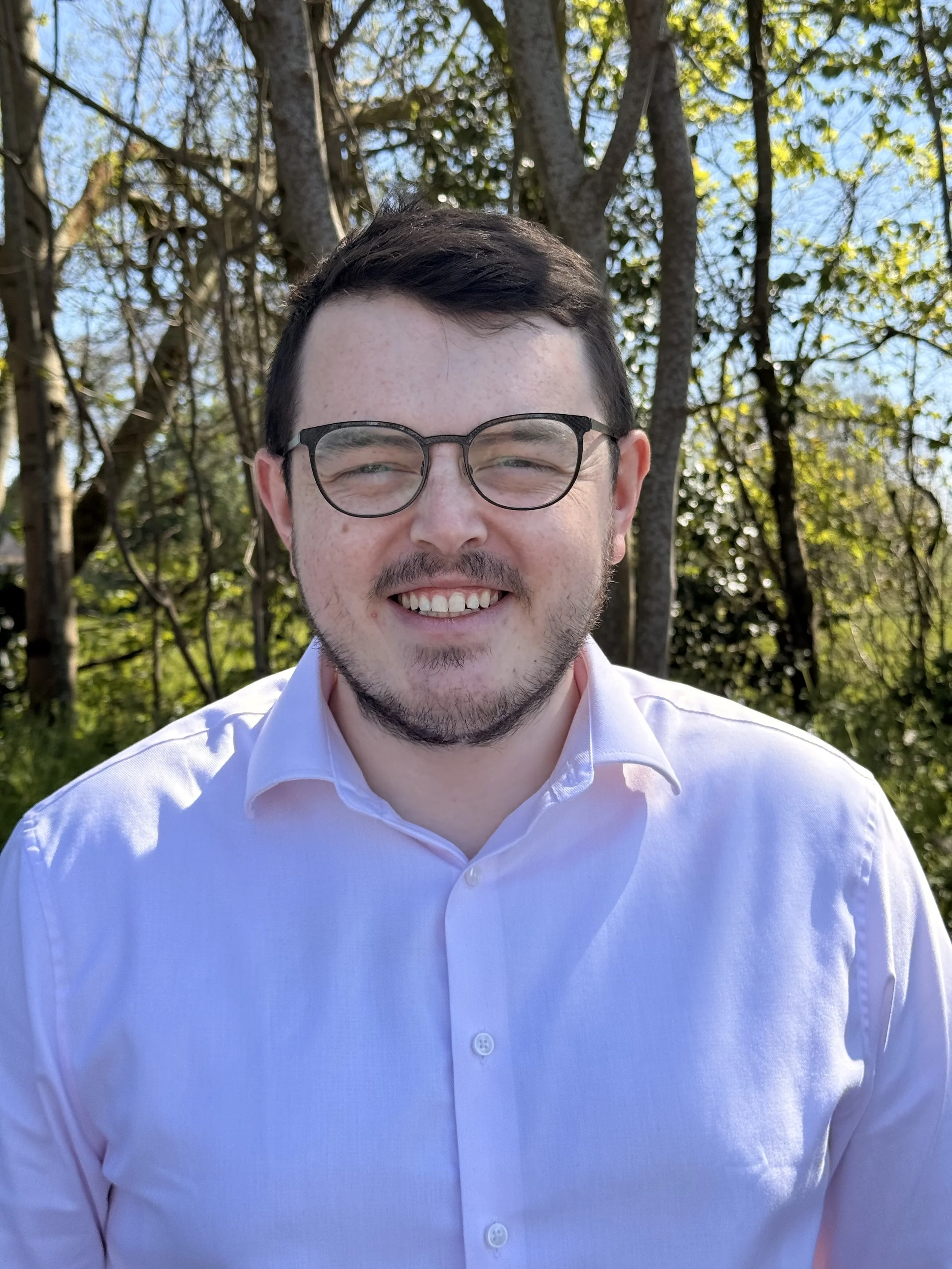 A man with short dark hair and glasses smiling outdoors with trees and a blue sky in the background, wearing a white button-up shirt.
