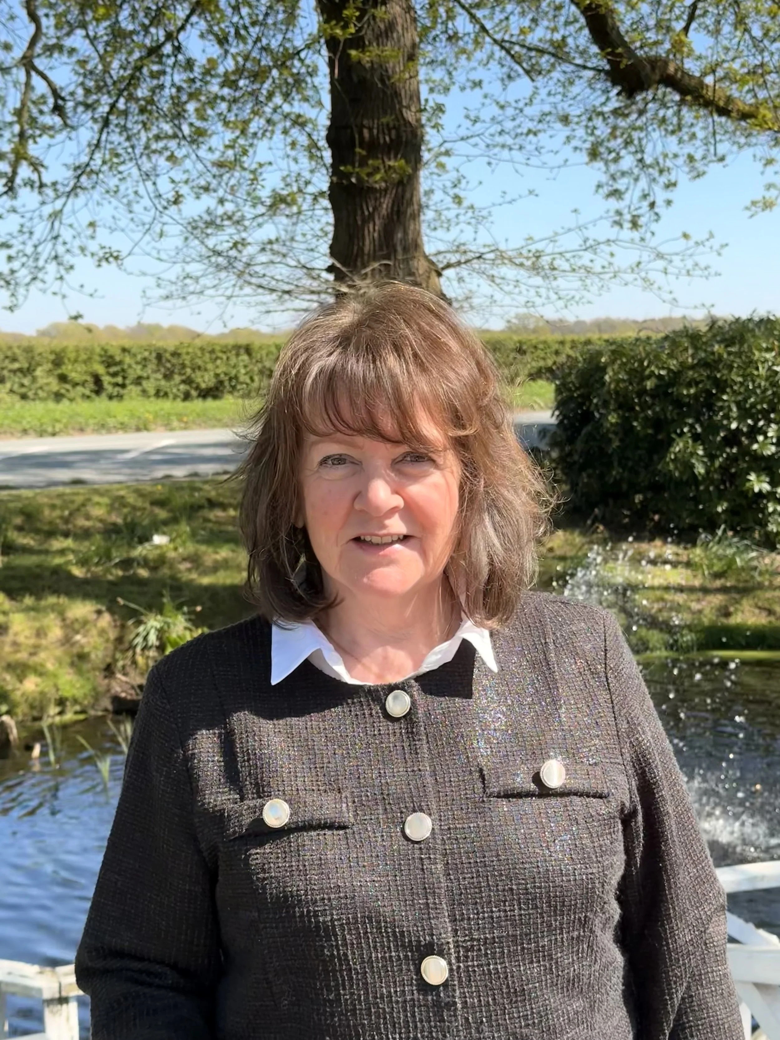 A woman with shoulder-length brown hair standing outdoors near a body of water, with trees and shrubs in the background, on a bright sunny day.