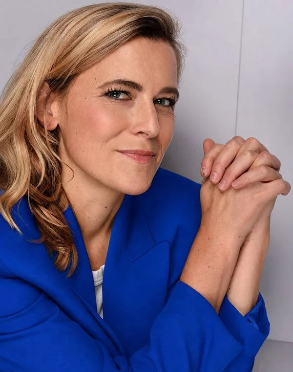 Eine Frau mit blonden Haaren, lächelnd, trägt eine blaue Blazer, sitzt vor einem weißen Hintergrund, die Hände sind gefaltet.