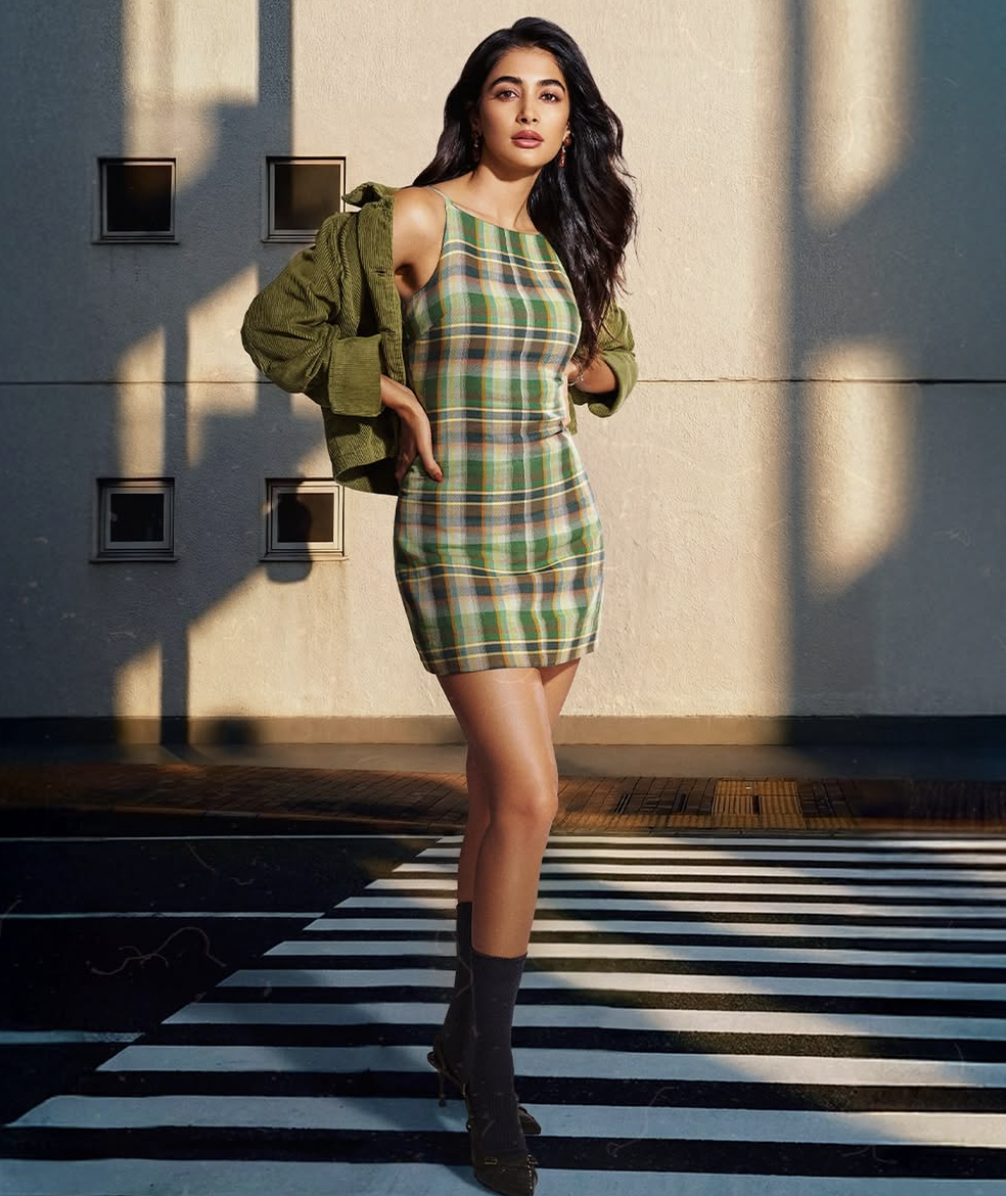 A young woman standing on a crosswalk in front of a building with sunlight and shadow effects. She is wearing a plaid dress, a green jacket, and black high-heeled boots.