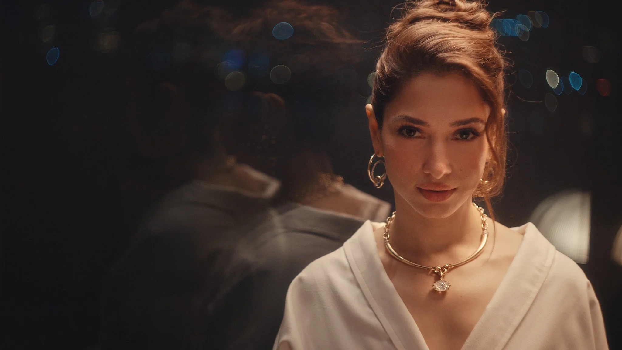 A woman with short curly red hair and gold jewelry looking at a person who is partially visible in the foreground, in a dimly lit setting with blurred colorful lights in the background.