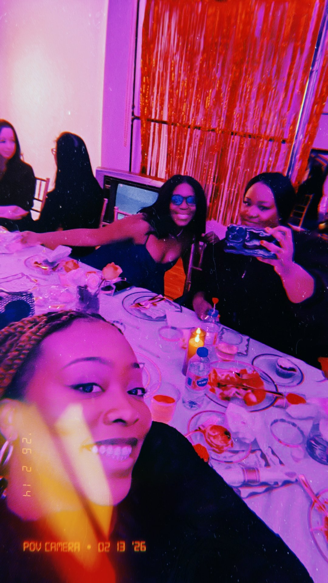 Group of women at a dinner party with a table full of food and drinks, taking a selfie in a restaurant or event space with colorful lighting and decorations.