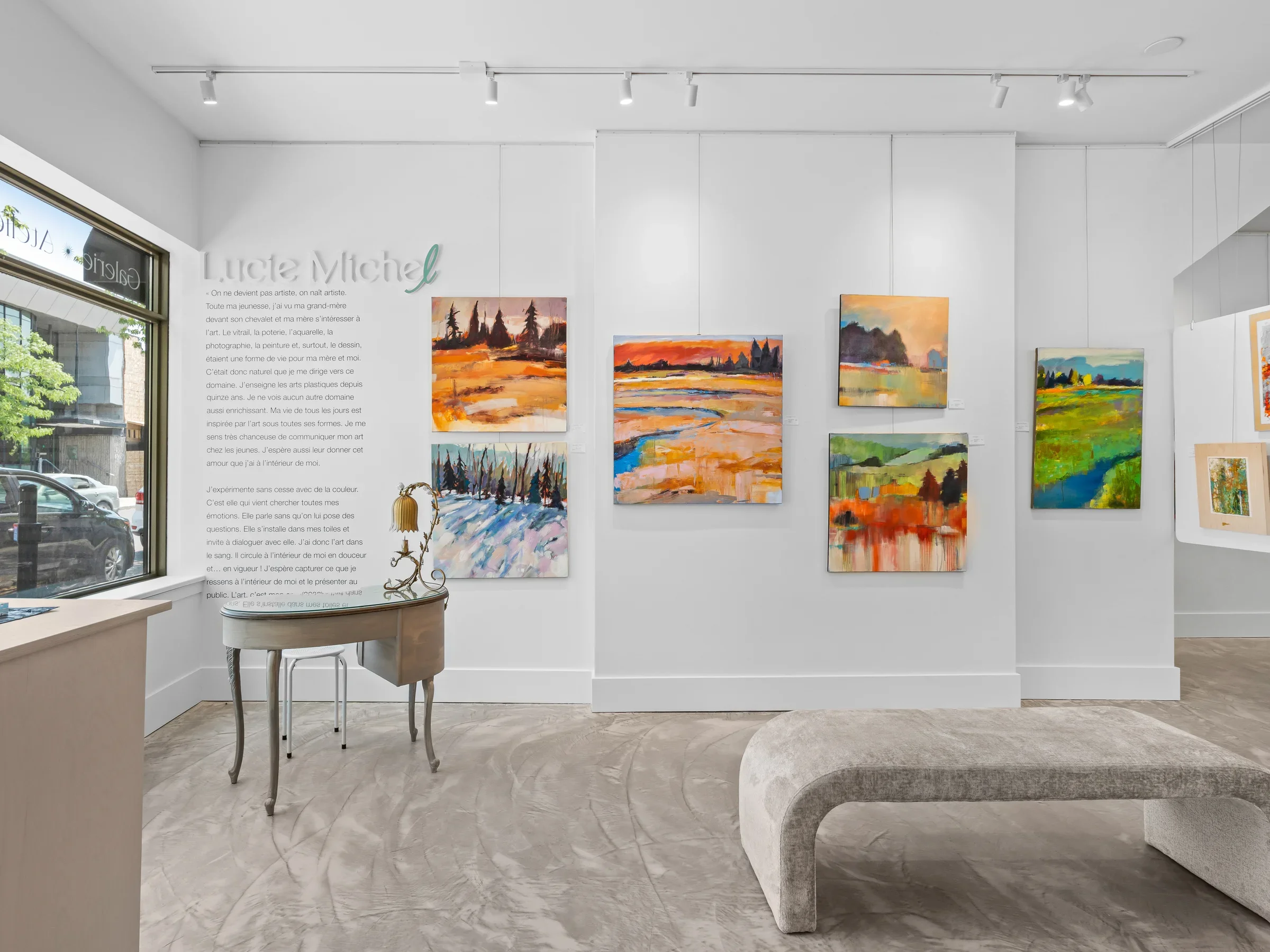 Art gallery with abstract landscape paintings on white walls, a window on the left, a decorative chair with a lamp on it, and a curved gray bench in the foreground.