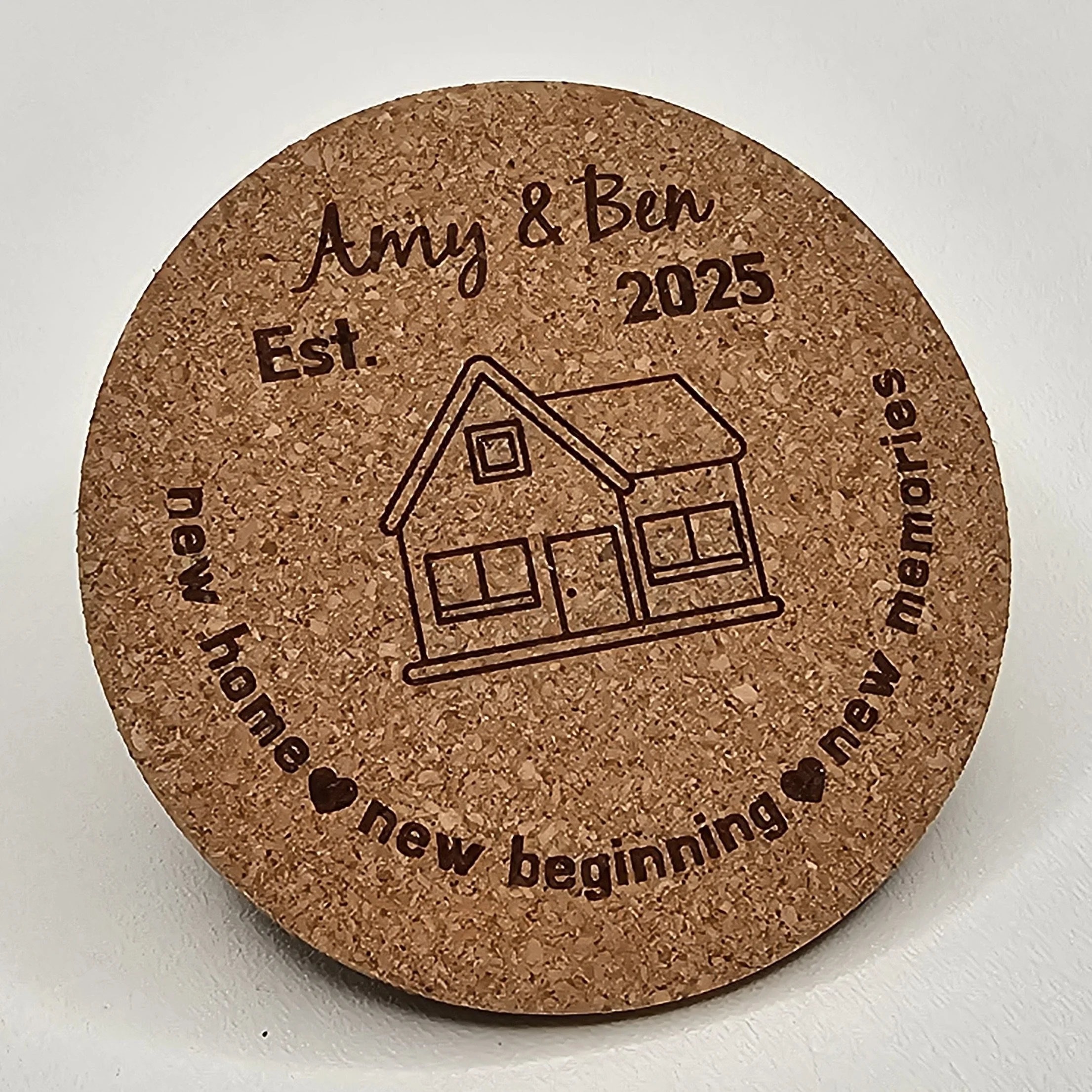 Round cork coaster engraved with a house drawing, text 'Amy & Ben 2025', 'Est. 2024', and a quote about home and new beginnings.