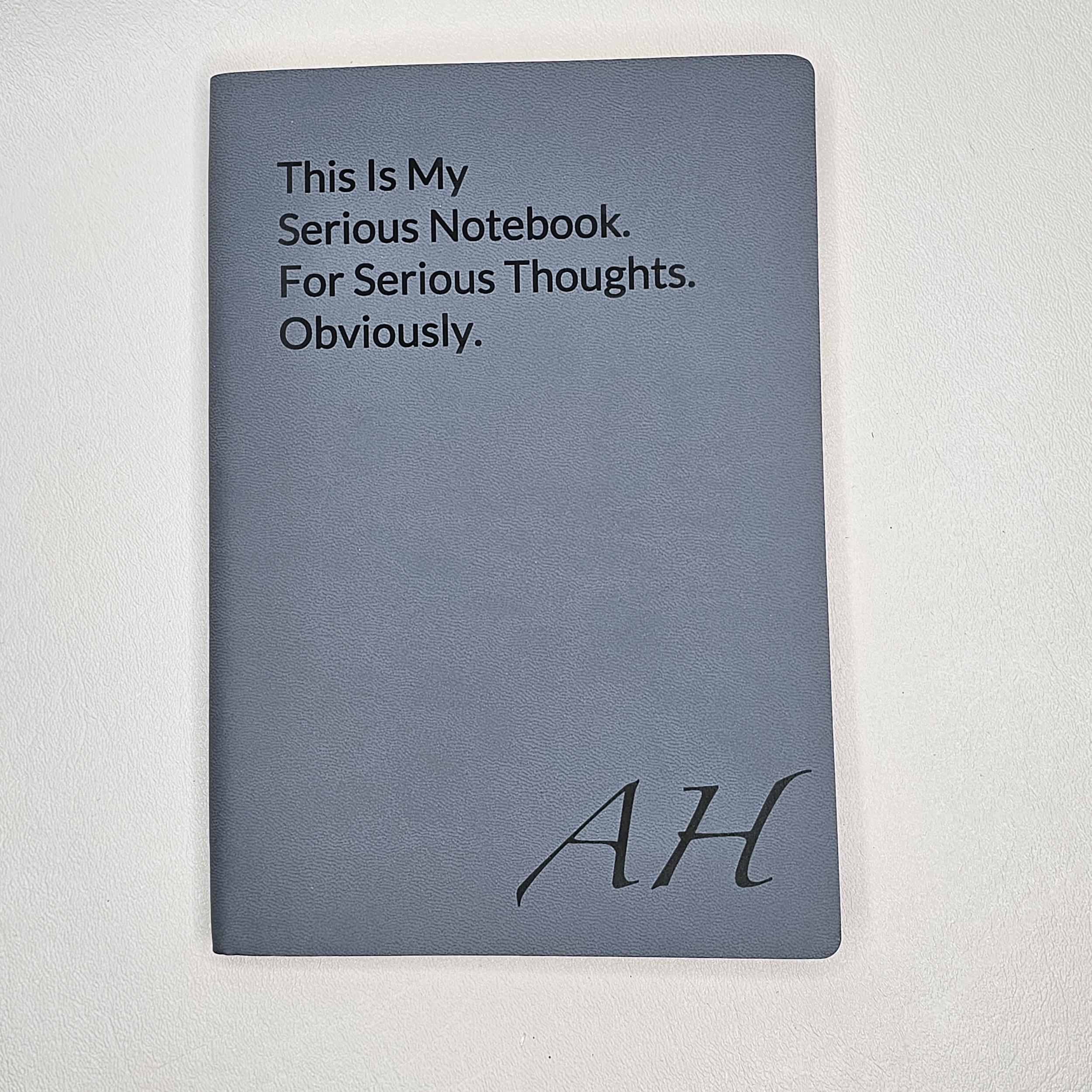 Gray notebook with the words 'This Is My Serious Notebook. For Serious Thoughts. Obviously.' written in black text, and the initials 'AH' in cursive at the bottom.