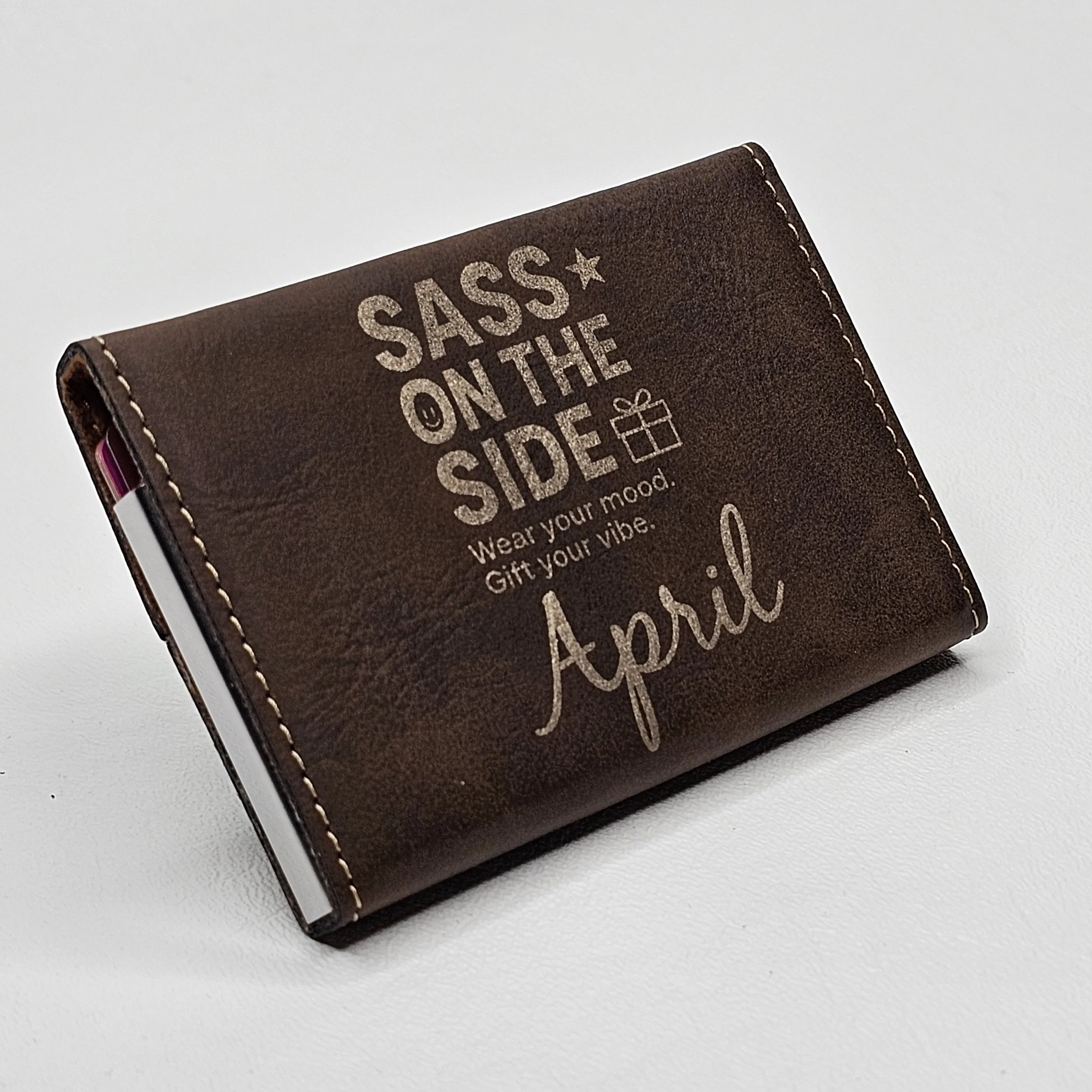 Brown wallet with printed text that reads "SASS ON THE SIDE, Wear your mood. Gift your vibe. Apri" and a small gift box icon.