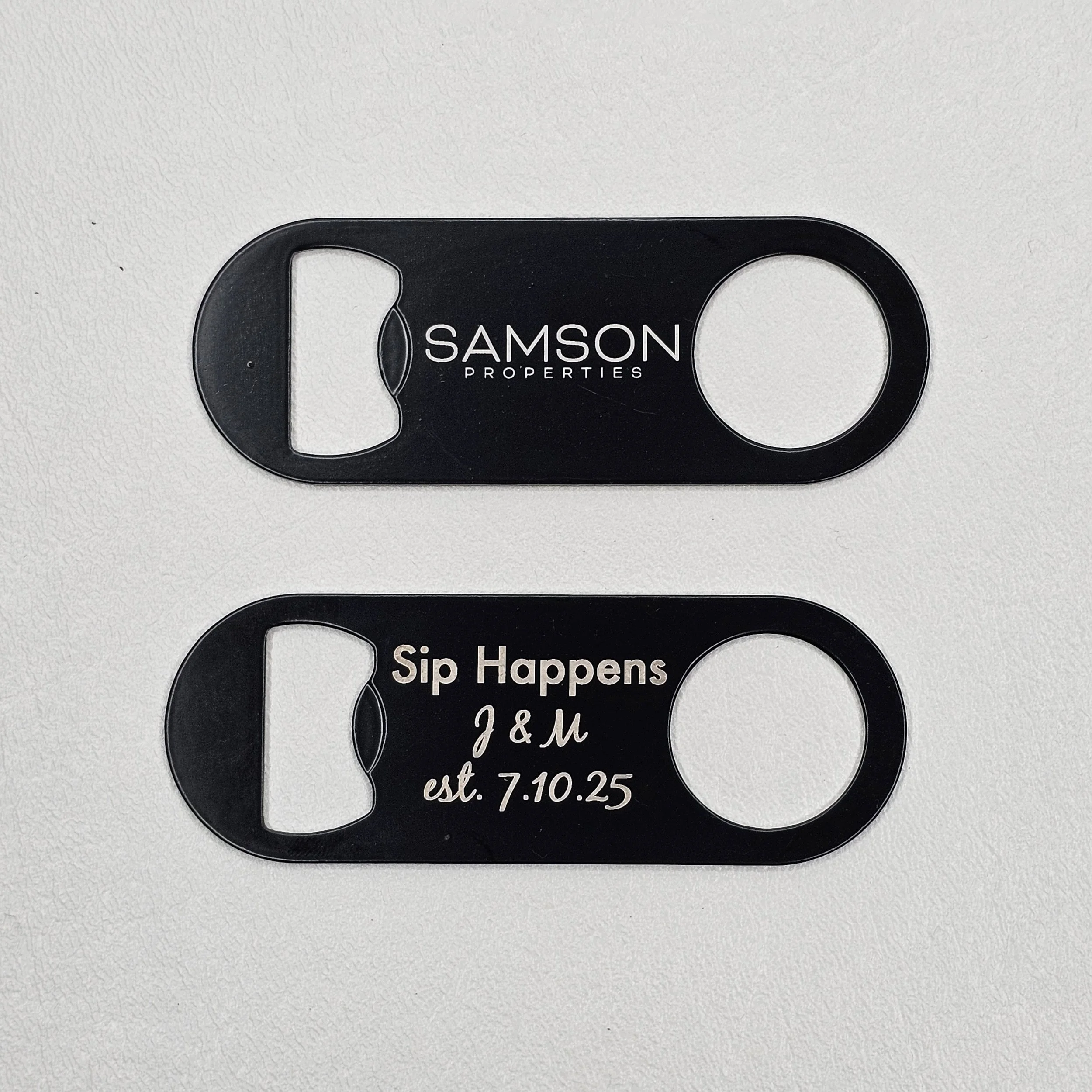 Two black drink coasters with openings for bottles, one with 'SAMSON PROPERTIES' logo and the other with 'Sip Happens J & M est. 7.10.25' written on them, resting on a white surface.