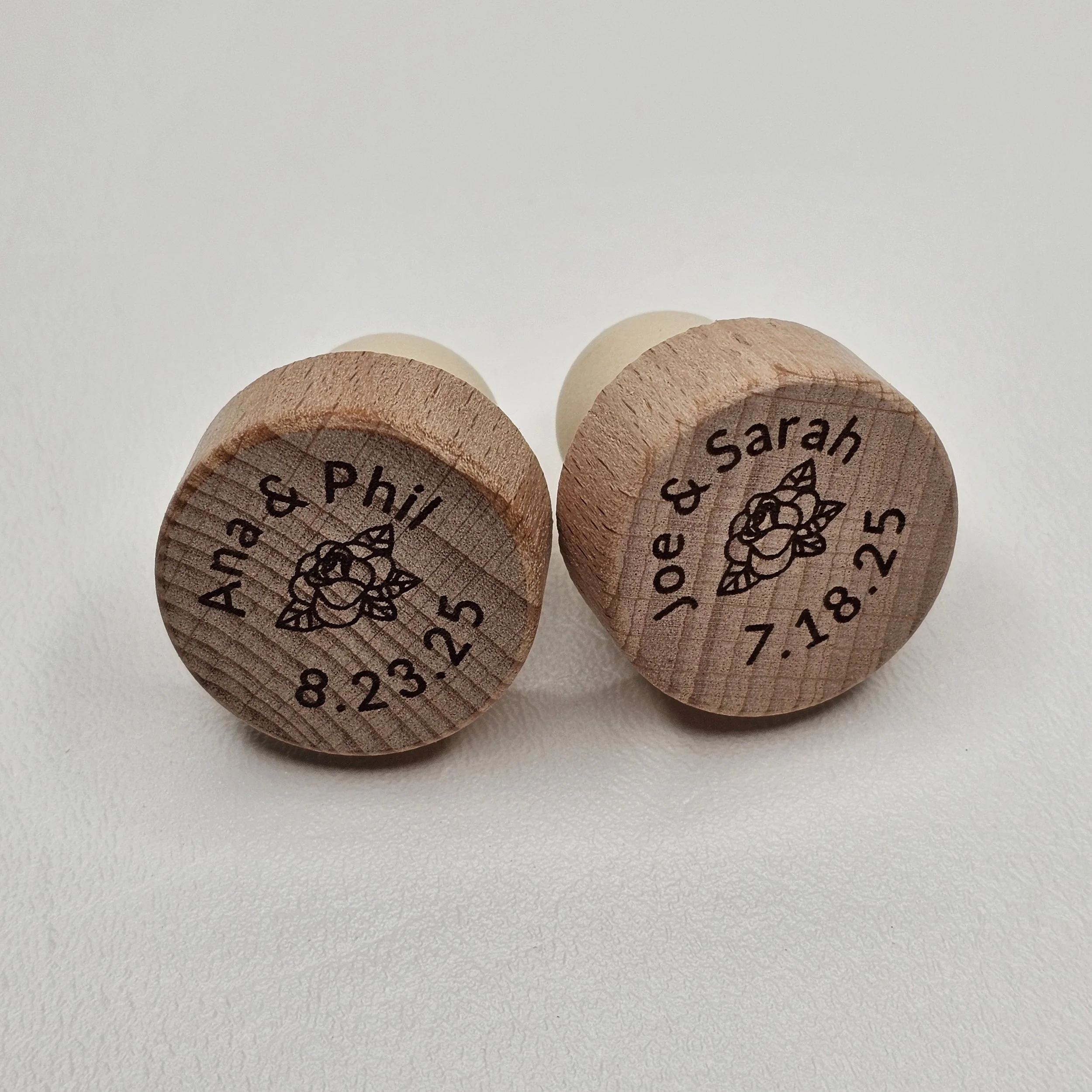 Two personalized wooden wedding stamps with dates and names, one labeled "Anna & Phil" with date "8.23.25", the other labeled "Joe & Sarah" with date "7.18.25", and a floral design in the center.