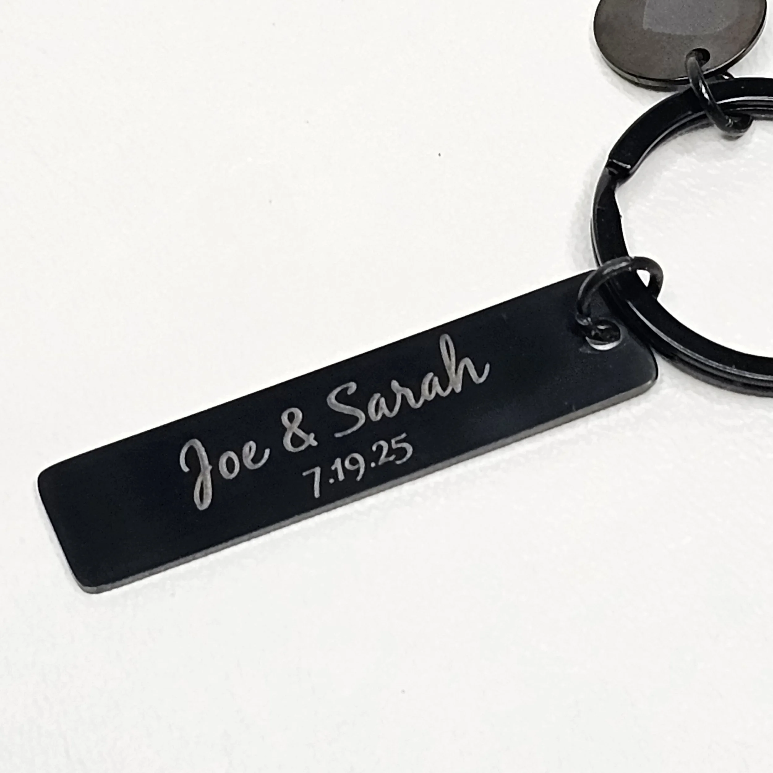 A black metal keychain with engraved text reading 'Joe & Sarah' and the date '7.19.25', attached to a larger key ring.