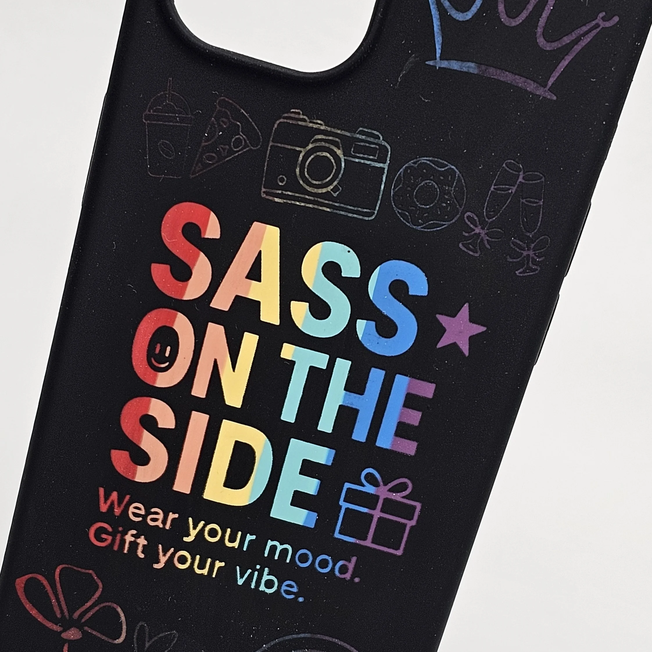 A black gift bag with colorful text and illustrations, reading 'SASS ON THE SIDE. Wear your mood. Gift your vibe.' and decorated with doodles of a drink, pizza, camera, donut, champagne glasses, star, gift box, and hearts.