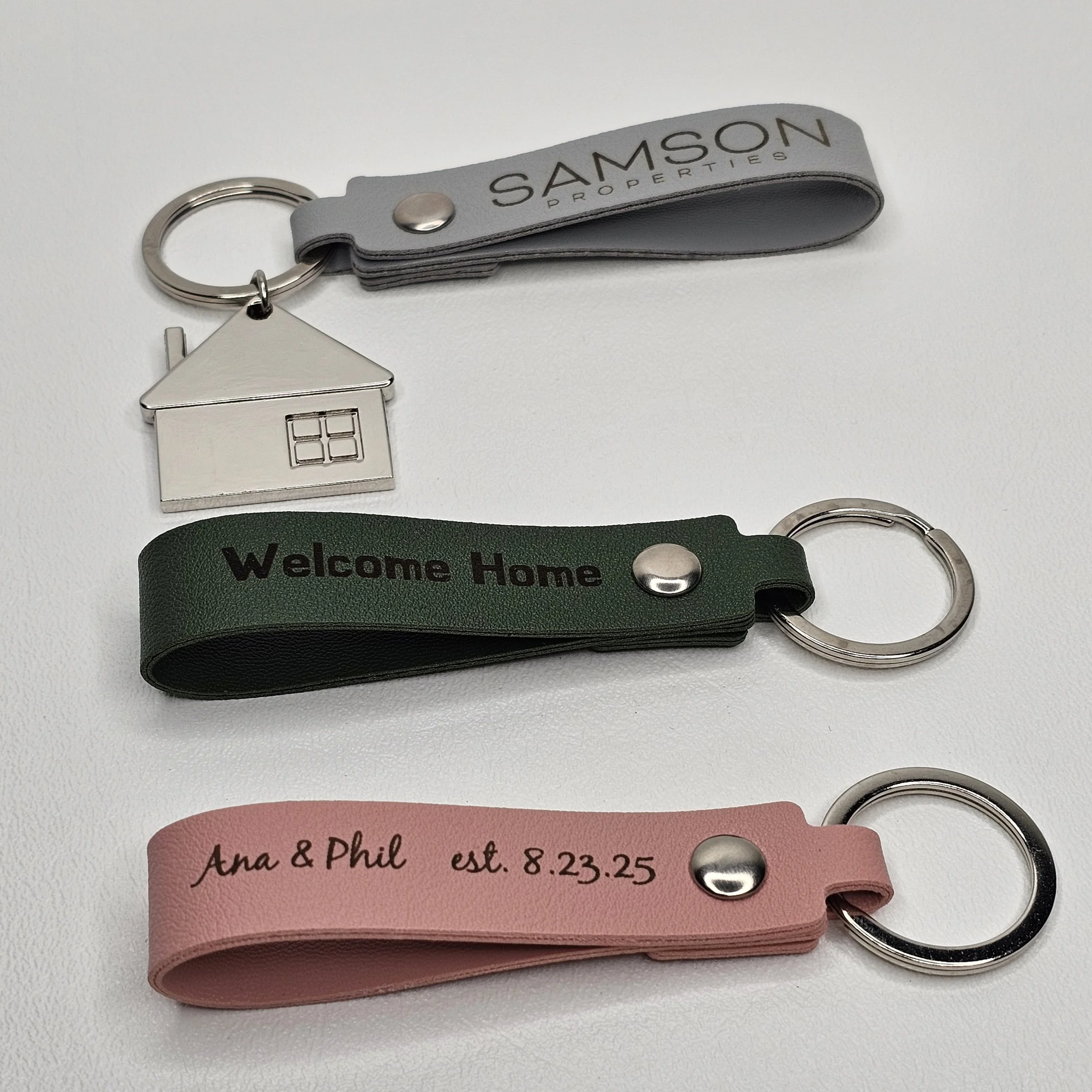 Three keychains with fabric straps; the first gray with 'SAMSON PROPERTIES', the second green with 'Welcome Home', and the third pink with 'Ana & Phil est. 8.23.25'; each attached to a metal ring.